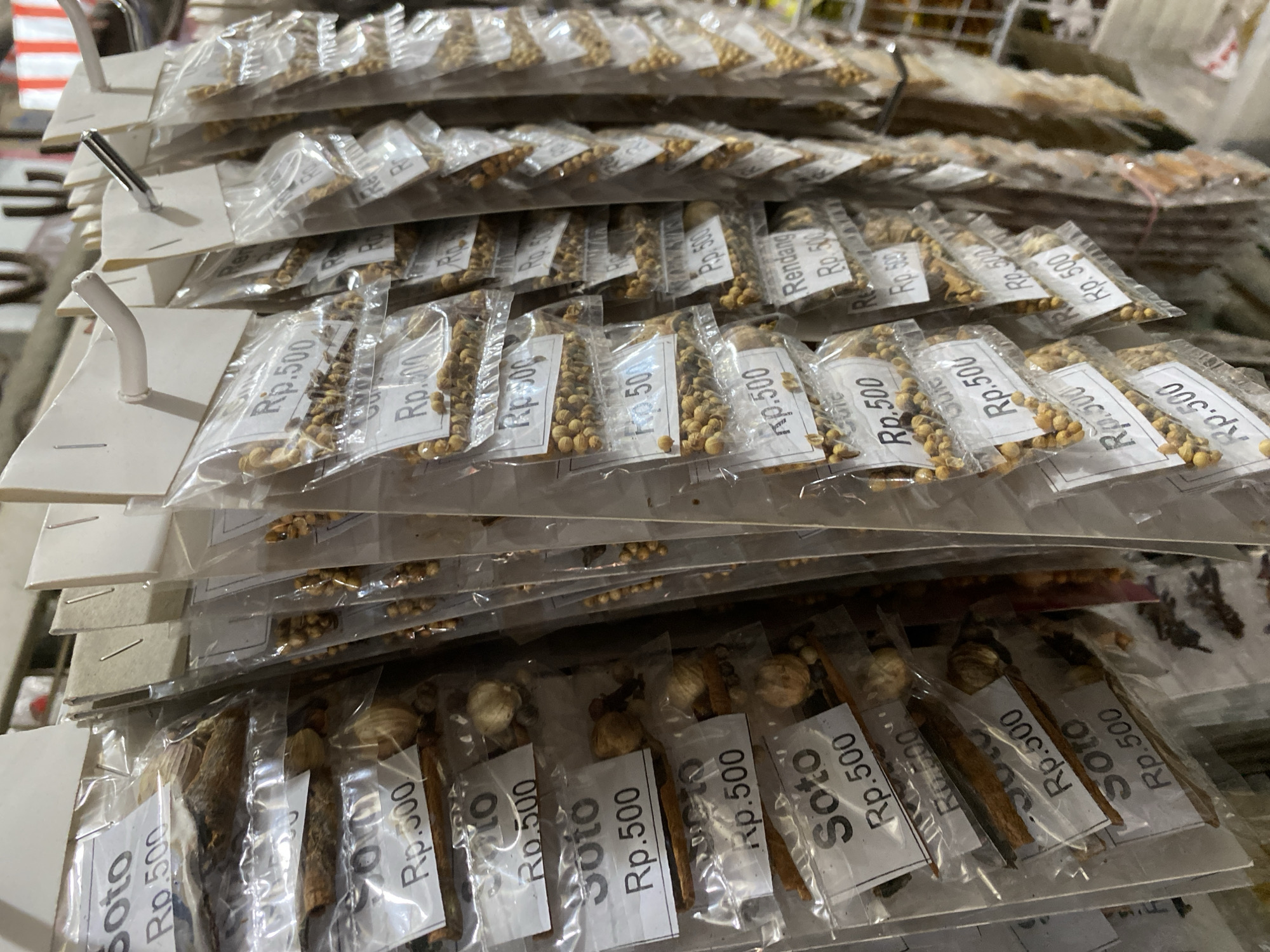 The image shows a retail display of various dried spices and herbs, individually packaged in clear plastic bags and hung on a rack. Each bag has a white label indicating the product name and price. The labels visible include "Soto" and "Rendang", both priced at "Rp.500". The spices appear to be in a market or shop setting, with other items and shelving visible in the background. The lighting suggests an indoor environment, possibly a traditional Indonesian market in Ambarketawang, Indonesia, given the context provided. There are no people visible in the frame, and the focus is solely on the products on display.