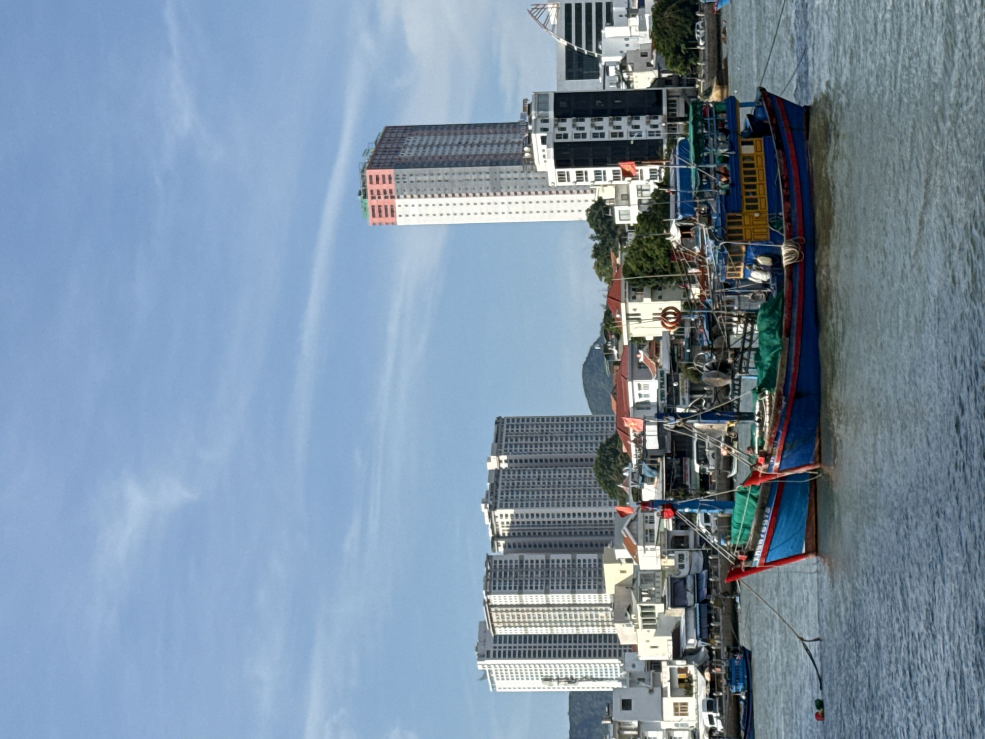 The image shows a waterfront scene in Bac Nha Trang Ward, Vietnam, under a clear blue sky with scattered wispy clouds. In the foreground, two vibrant blue and red fishing boats are docked. The boats have green netting and are tied together with ropes. One boat has a yellow cabin and several figures are visible on deck. In the middle ground, a variety of buildings line the waterfront, including residential structures, shops, and businesses. Several red flags are visible on poles atop some of these buildings. In the background, a cluster of tall white apartment buildings dominates the skyline, with a distinctive tall, pink and white skyscraper under construction to the right. Green hills can be seen behind the buildings. The water is a calm, rippling blue.