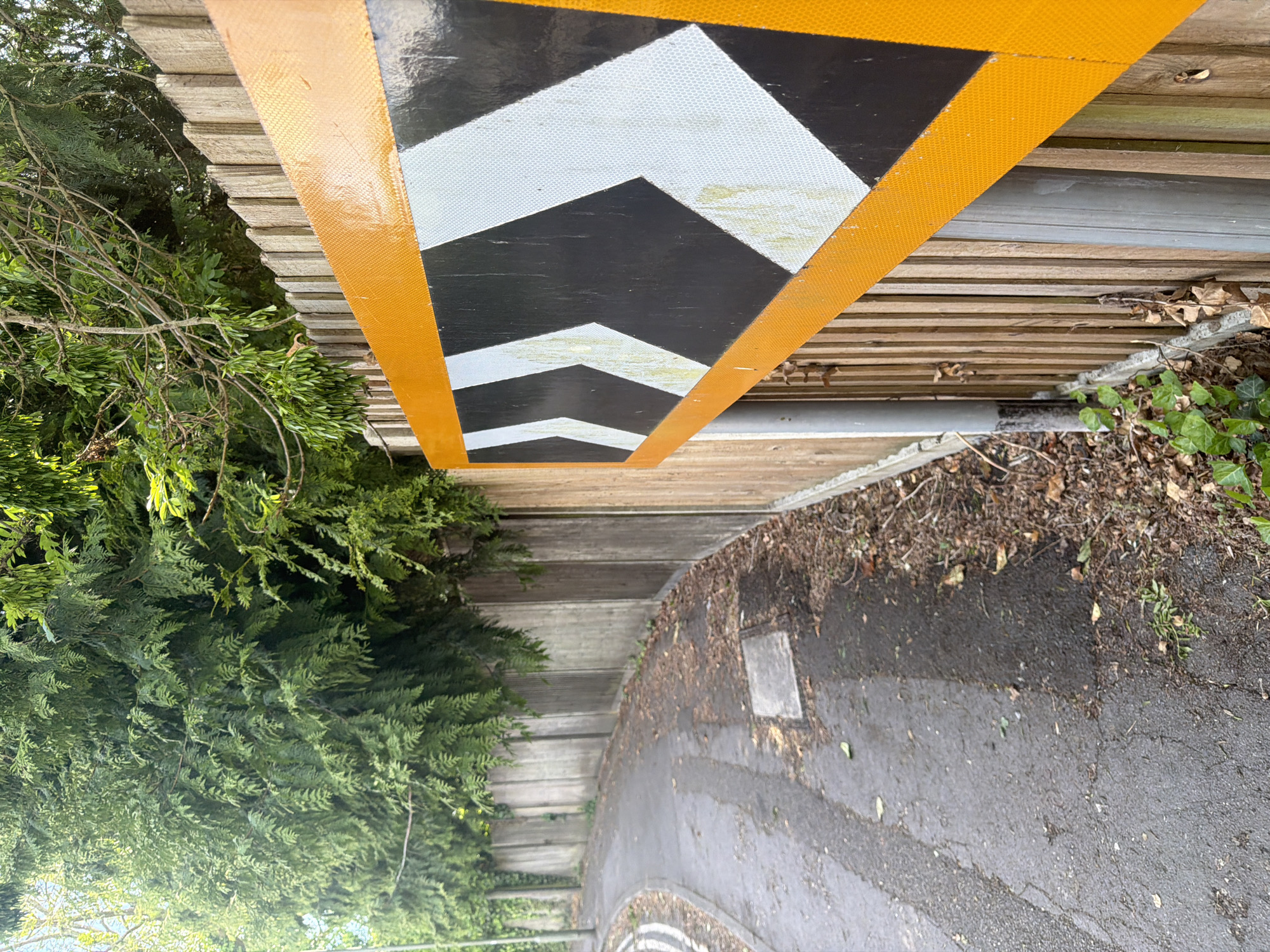 The image depicts a street scene in Guildford, United Kingdom. A section of roadway is visible, featuring a black asphalt surface. A triangular road sign with reflective white and black directional arrows, bordered in orange, is affixed to a wooden structure. A section of the wooden structure is above a concrete wall, with shrubbery and trees bordering the other side. The scene appears to be in an outdoor environment with natural lighting. No people are visible in the image.