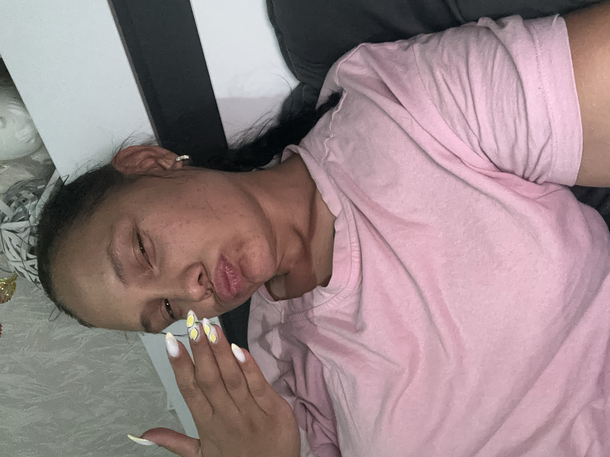 An individual, appearing to be a woman, is depicted in an indoor setting. She has medium-to-dark skin, dark hair pulled back, and visible freckles on her face. Her left eye is partially closed in a wink, and her lips are pursed into a prominent pout. She is wearing a light pink, short-sleeved, crew-neck t-shirt, exhibiting wrinkles and fabric texture. A small, silver-toned hoop earring, possibly with embedded stones, is visible on her right ear. Her left hand is raised, with fingers slightly splayed, partially obscuring her lower face. Her fingernails are long, almond-shaped, and painted white; the middle and ring fingers feature yellow lemon slice designs. A long, dark braid or ponytail rests over her right shoulder. The background consists of a light grey textured wall, a white panel with a horizontal black stripe, and a dark grey upholstered surface, likely a headboard or sofa. Partially visible in the upper left background are some gold-colored decorative objects. The indoor environment features generic decor and offers no specific visual identifiers to confirm its location as Sofia, Bulgaria.