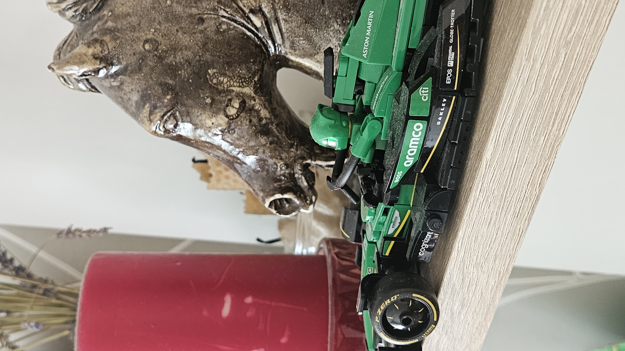 A Lego Formula 1 race car, green and black with yellow accents and sponsor logos, is positioned on a light-colored wooden shelf. The car features the "Aston Martin" logo on its rear wing. To the left of the car is a tall, vibrant red candle. Behind the car and to the right is a textured, dark brown ceramic horse head statue. In the background, out of focus, are dried lavender stems and other indistinct objects. The scene is indoors, illuminated by natural light. The image does not provide any information or clues about the city of Bratislava, Slovakia.