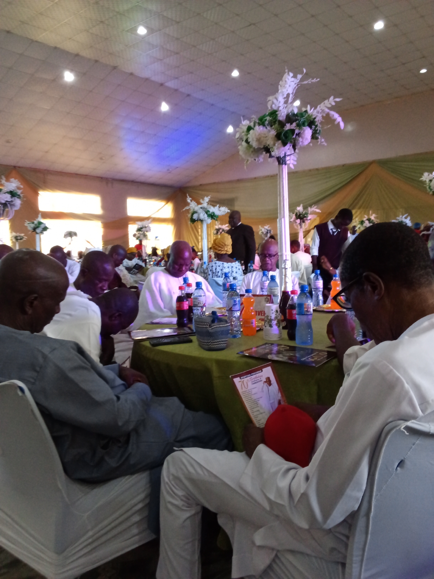 The image depicts an indoor gathering, likely a reception or celebration, set in a venue in Barden Yana, Nigeria. In the foreground, several men are seated at tables draped with green tablecloths. One man, in the immediate foreground and facing away from the camera, wears a white garment and is looking down at a red cap and a booklet titled "RECEPTION PROGRAM 70th Birthday". Another man to his left wears a grey traditional outfit and is also looking down. At the table in the center, several men are seated, some looking down at their laps or the table, while others are engaged in conversation. Bottles of drinks, including Coca-Cola and water, are visible on the table. In the background, more guests are present, some standing and conversing. The venue appears to be decorated with floral arrangements and draped fabric, with lighting from ceiling fixtures. The overall atmosphere suggests a formal or semi-formal event.