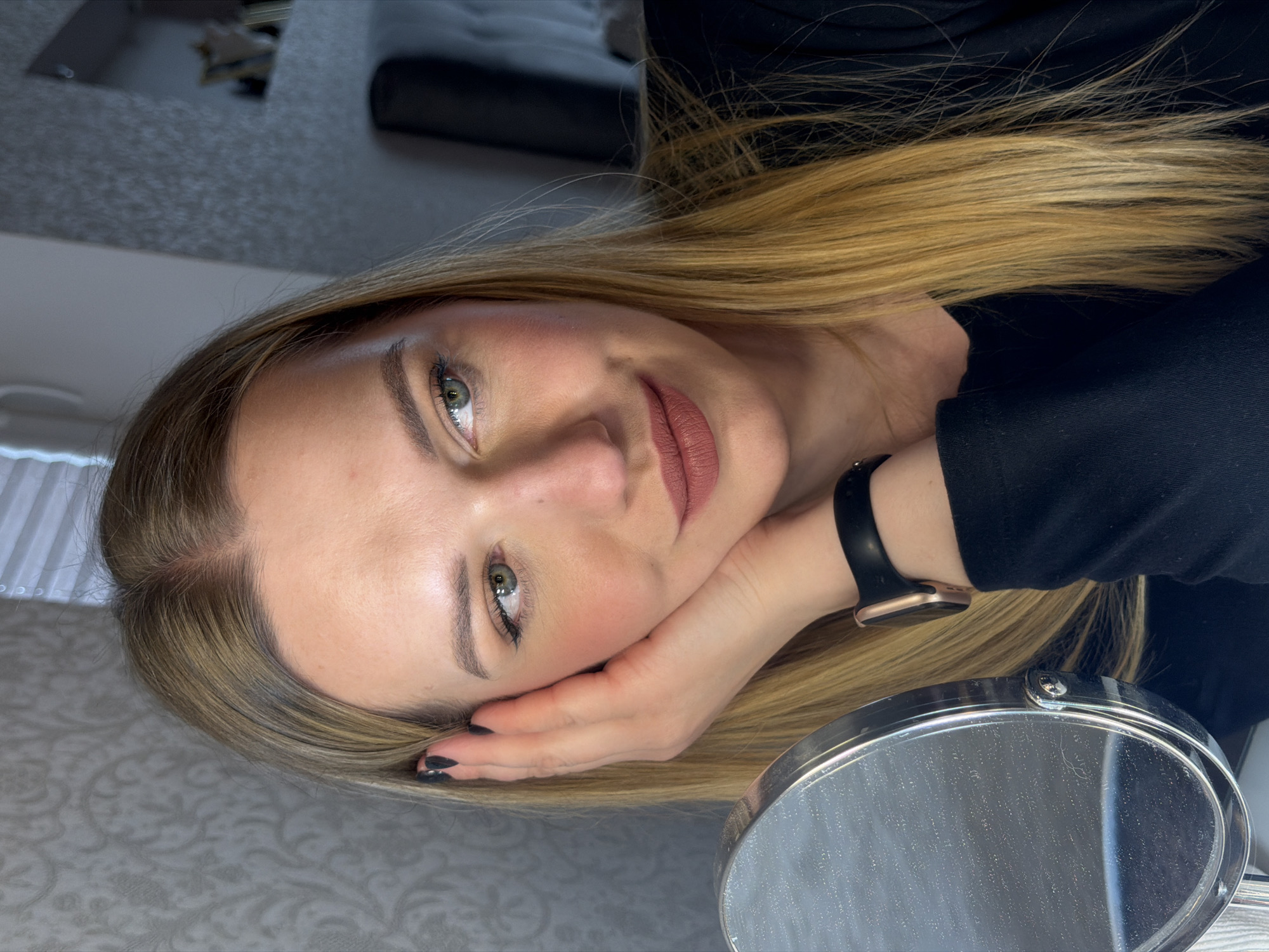 A fair-skinned woman with light blonde hair is shown from a close-up, angled perspective. She rests her head on her left hand, displaying a black smartwatch on her left wrist. Her nails are painted black. Her facial makeup includes defined eyebrows, eyeliner, blush, and muted pink lipstick. A small, round, metal-framed mirror is positioned near her face. The background includes indistinct shapes and surfaces. The exact location within the city of Dreiliņi, Latvia is not discernible from the image.