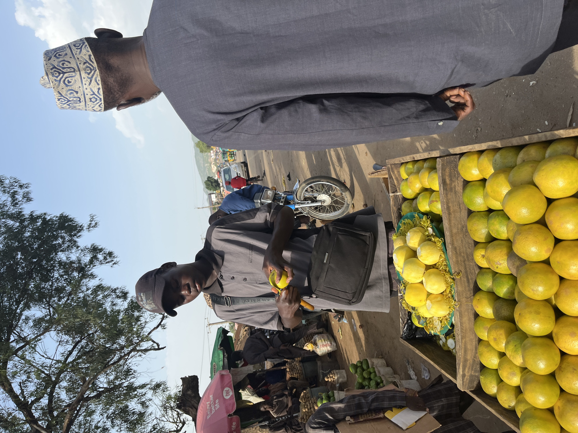 In Rafin Par, Nigeria, a young male vendor wearing a grey polo shirt and a baseball cap is peeling a bright yellow fruit, likely a mango or a lemon, with a small knife. He is standing behind a wooden crate filled with vibrant yellow and green citrus fruits, possibly oranges, and other produce. Beside him, a dark-colored messenger bag hangs from his shoulder. Facing him, an older male, dressed in a dark blue tunic and a white and blue patterned cap, is partially visible from the back, his left hand resting on his hip. The market scene unfolds on a dirt ground, with a blue motorcycle parked nearby. In the background, a bustling street is visible with cars, a tricycle, and other market stalls under a clear blue sky with scattered clouds, shaded by the branches of a large tree.