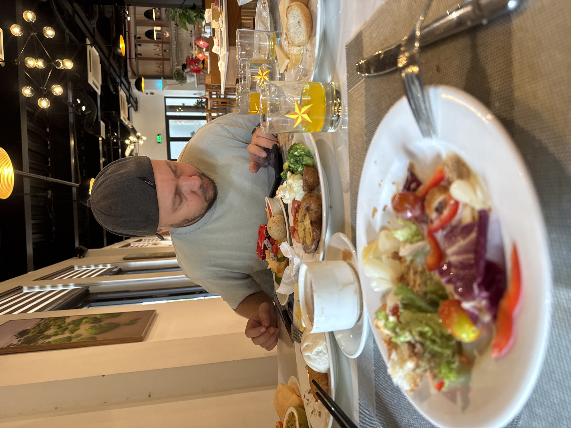 A man wearing a gray cap and a light green t-shirt is seated at a table laden with food and beverages. He appears to be looking at his phone. The table includes plates of salad, various dishes, and glasses of orange juice. A white cup is also on the table. Utensils are placed next to the plates.
In the background, through a doorway, a restaurant setting is visible with people dining. Lights hang from the ceiling, and artwork is displayed on the wall. The scene is set in what appears to be a restaurant in the city of Đà Nẵng, Vietnam, as indicated by the small flags placed on tables. The interior is well-lit.