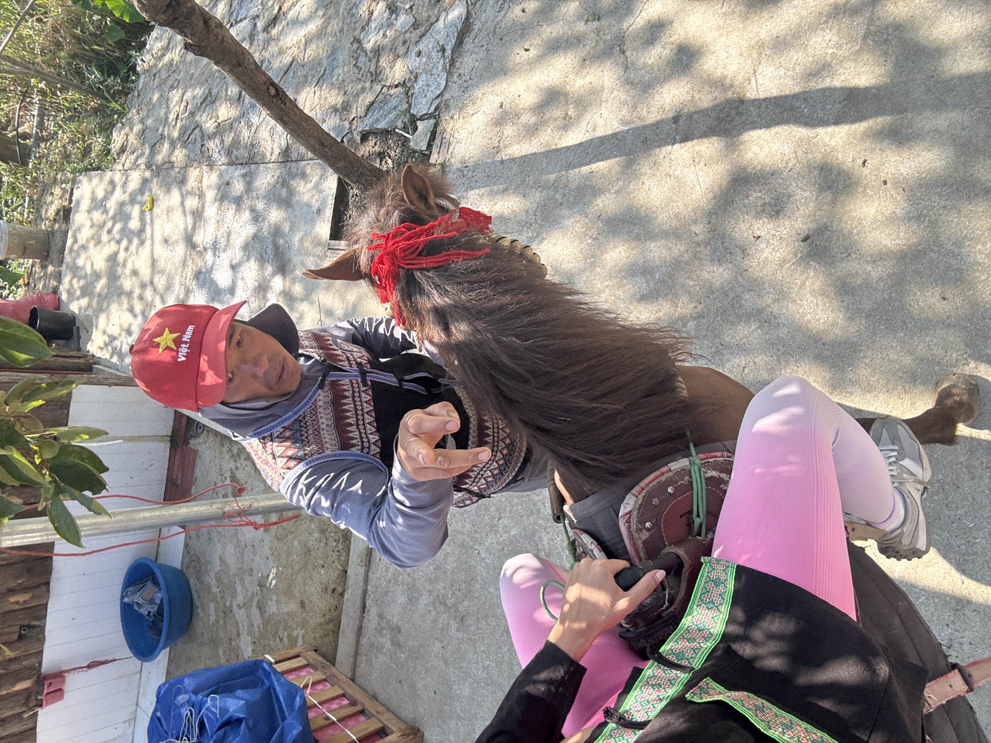 A man in a red "Việt Nam" hat is shown in an eye-level shot, standing beside a horse with a red head ornament. The horse is saddled, and someone wearing pink leggings and black clothing is sitting on the horse. The location is likely in Bắc Hà, Vietnam, though it is not definitively shown. The setting appears to be an outdoor area with concrete paving and some vegetation.