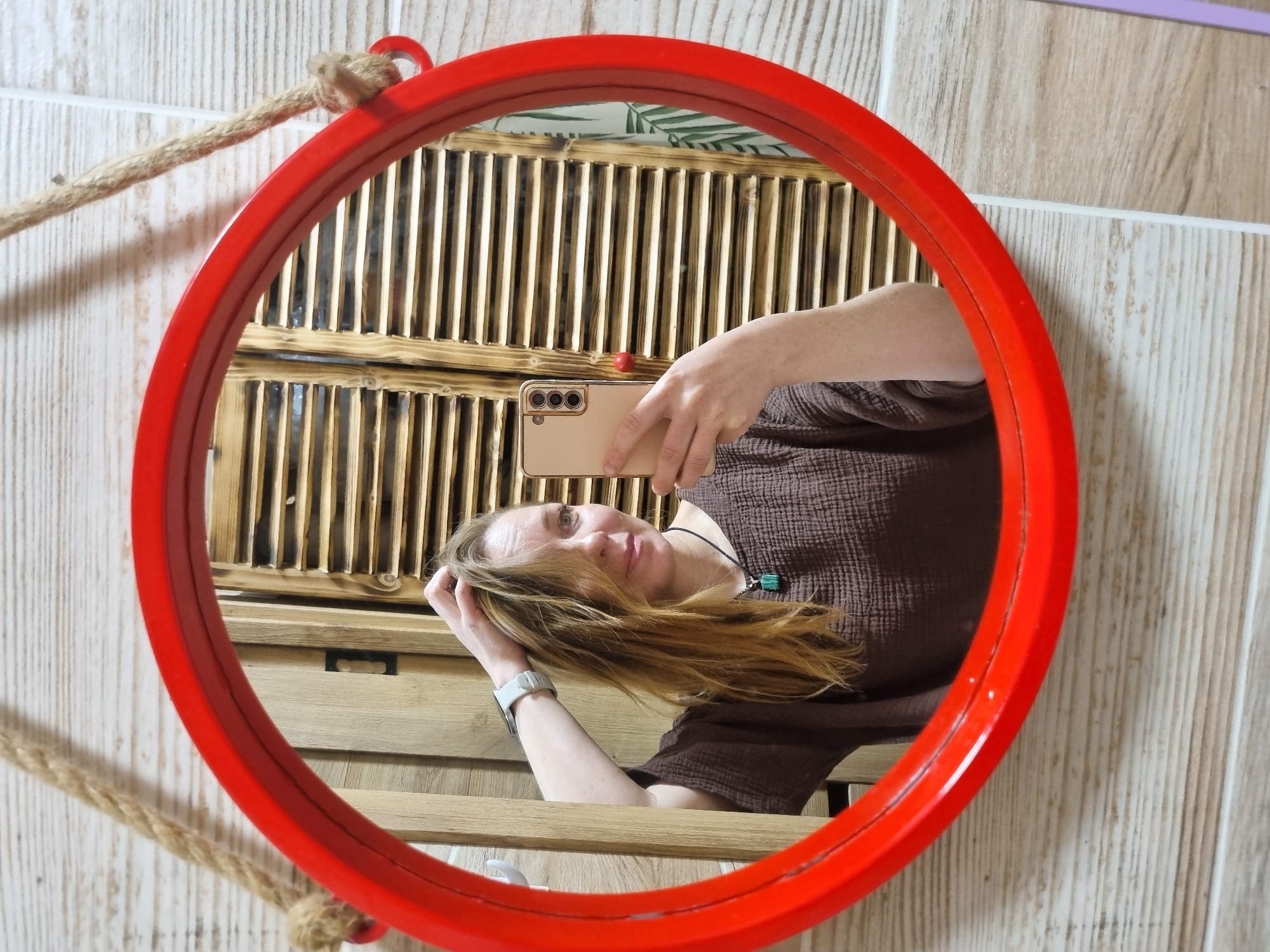 A person is taking a selfie in a round, red-framed mirror. The mirror hangs on a wall of light-colored, vertical tiles with a brown rope. Reflected in the mirror is a woman wearing a brown, short-sleeved shirt, holding a tan-colored smartphone. Behind her, also reflected, is a wooden slat wall above a wooden shelf or cabinet. A green plant appears on top of the slat wall. The location is indicated to be Chaikovsky, Russia, though no specific features of the location are identifiable in the image.