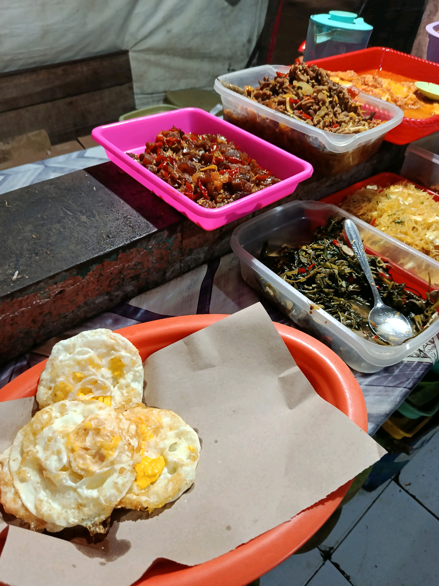 In the city of Kudus, Indonesia, a close-up, eye-level shot reveals a street food stall setup. The scene is dimly lit, likely at night, with the overhead lighting creating shadows. In the foreground, an orange plastic plate holds three fried eggs served on brown paper. Behind this, a variety of dishes are presented in plastic containers. To the left, a pink plastic tray contains a stir-fried dish with red chilies and what appears to be meat. Next to it, a clear plastic container holds a dark green, leafy stir-fried vegetable dish, with a silver spoon resting inside. Further back, a red plastic tray displays noodles, and another clear plastic container holds a dish with shredded ingredients. A blue plastic jug is visible in the background. The surfaces are a mix of patterned fabric, wood, and tiled flooring, indicating an outdoor or open-air setting typical of Indonesian street food vendors. There are no people visible in the frame.