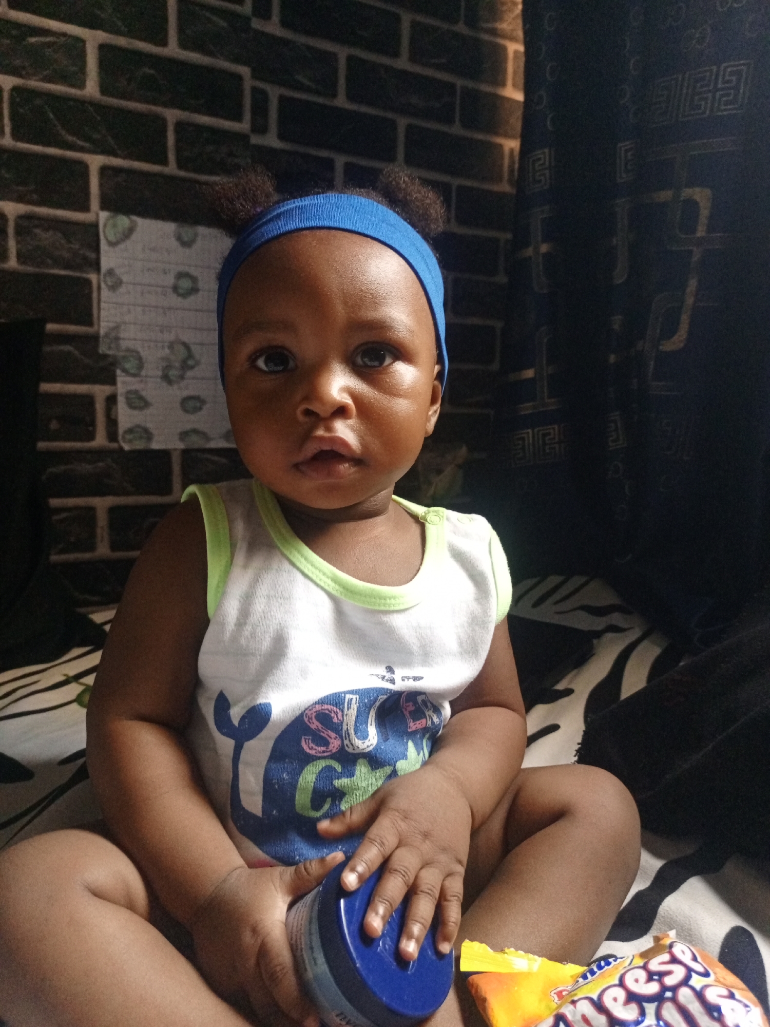 A baby with dark skin and dark eyes sits upright, looking directly at the camera. The baby wears a blue headband and a white sleeveless top with light green trim. On the front of the top, there is a graphic design featuring a blue whale or fish and partially visible text that appears to read "SU... G...".
The baby is holding a blue container, which appears to be a Nivea cream jar, with the text "NIV..." visible. To the baby's right, an orange and yellow snack bag is visible, with the word "Cheeseballs" or "Cheesepuffs" partially printed on it.
The baby is positioned on what appears to be a bed or blanket with a black and white striped pattern, possibly resembling a zebra print. In the background, there is a wall covered with dark, brick-patterned wallpaper or tiles. To the right of the baby's head, a dark patterned curtain or fabric with geometric shapes, possibly "GG" patterns, is visible. Behind the baby, attached to the brick wall, is a piece of white paper or fabric with light green circular patterns. The lighting suggests an indoor setting, possibly with indirect natural light or artificial light, creating a somewhat dim but clear view of the baby. The baby's expression is neutral and observant.