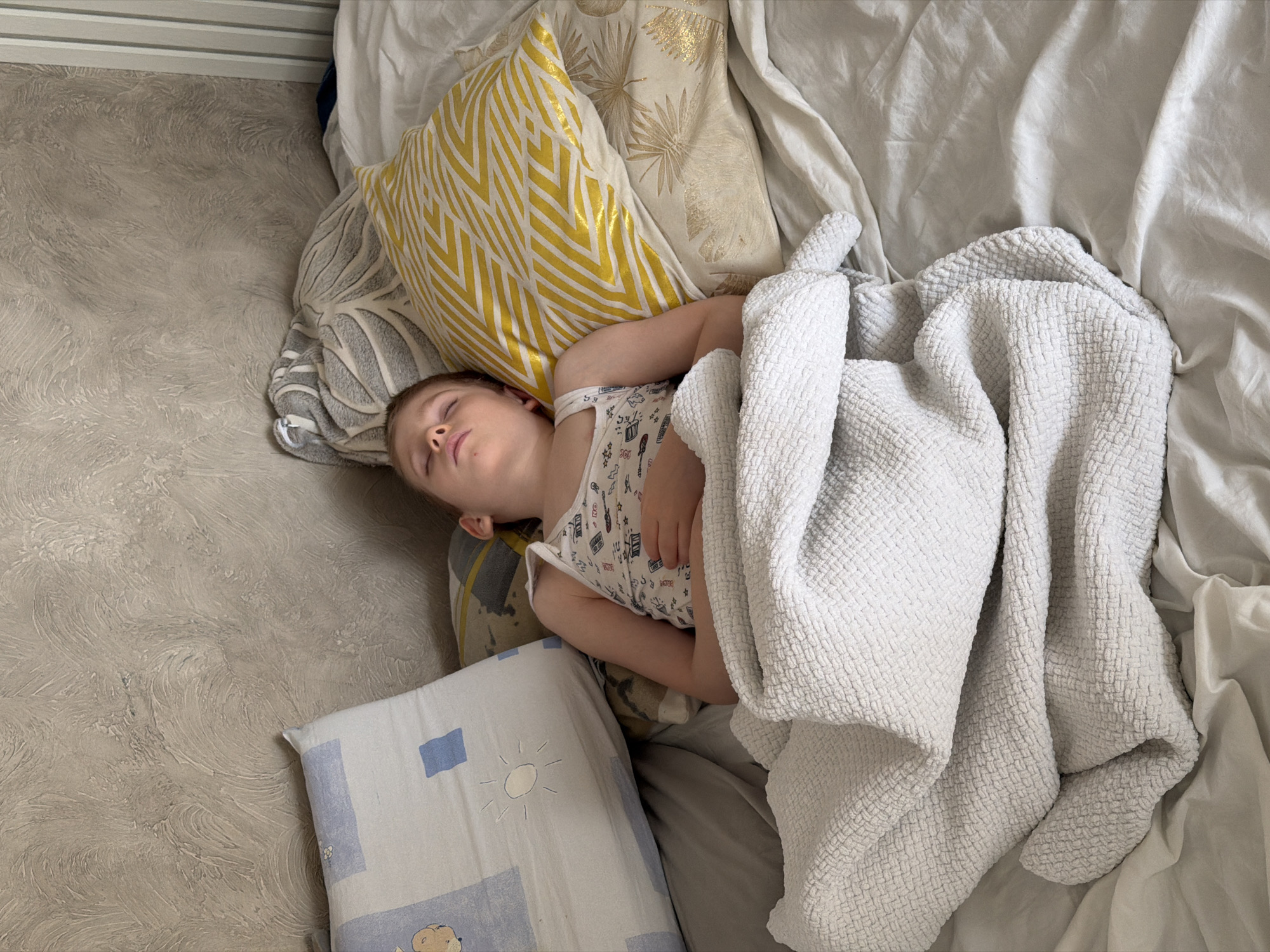 A child is sleeping on a bed with several pillows and a white blanket in a room in Rossiyskiy, Russia. The child is lying on their side, wearing a white tank top, and has their eyes closed. Several pillows surround the child, one with yellow and white chevron patterns, another with a gold design, and another with a blue and white design. The bed is covered with white sheets and a textured white blanket partially covers the child. The floor next to the bed is a light beige color.