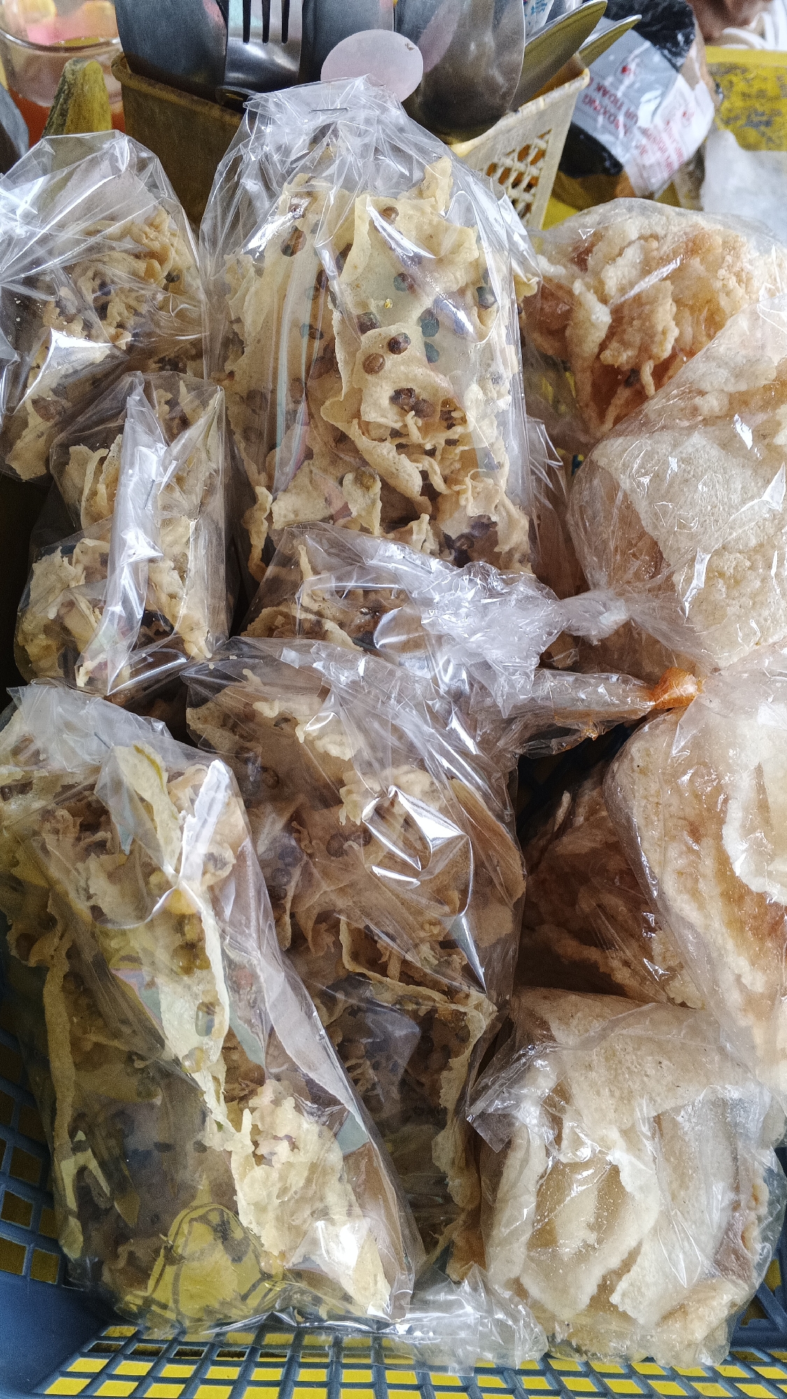 The image shows a close-up, overhead view of a blue plastic basket filled with several plastic bags of Indonesian snacks, possibly from a market stall in Yogyakarta, Indonesia. The snacks appear to be crispy and airy, with some bags containing thin, patterned crackers with visible dark specks, likely nuts or seeds, and others containing lighter, puffier chips. The plastic packaging is translucent, allowing the texture and color of the snacks to be seen. The basket's grid pattern is visible at the bottom, with a yellow hue showing through the blue plastic. In the background, out of focus, are hints of other items, including silverware and potentially other food items in packaging, suggesting a vendor's stall.