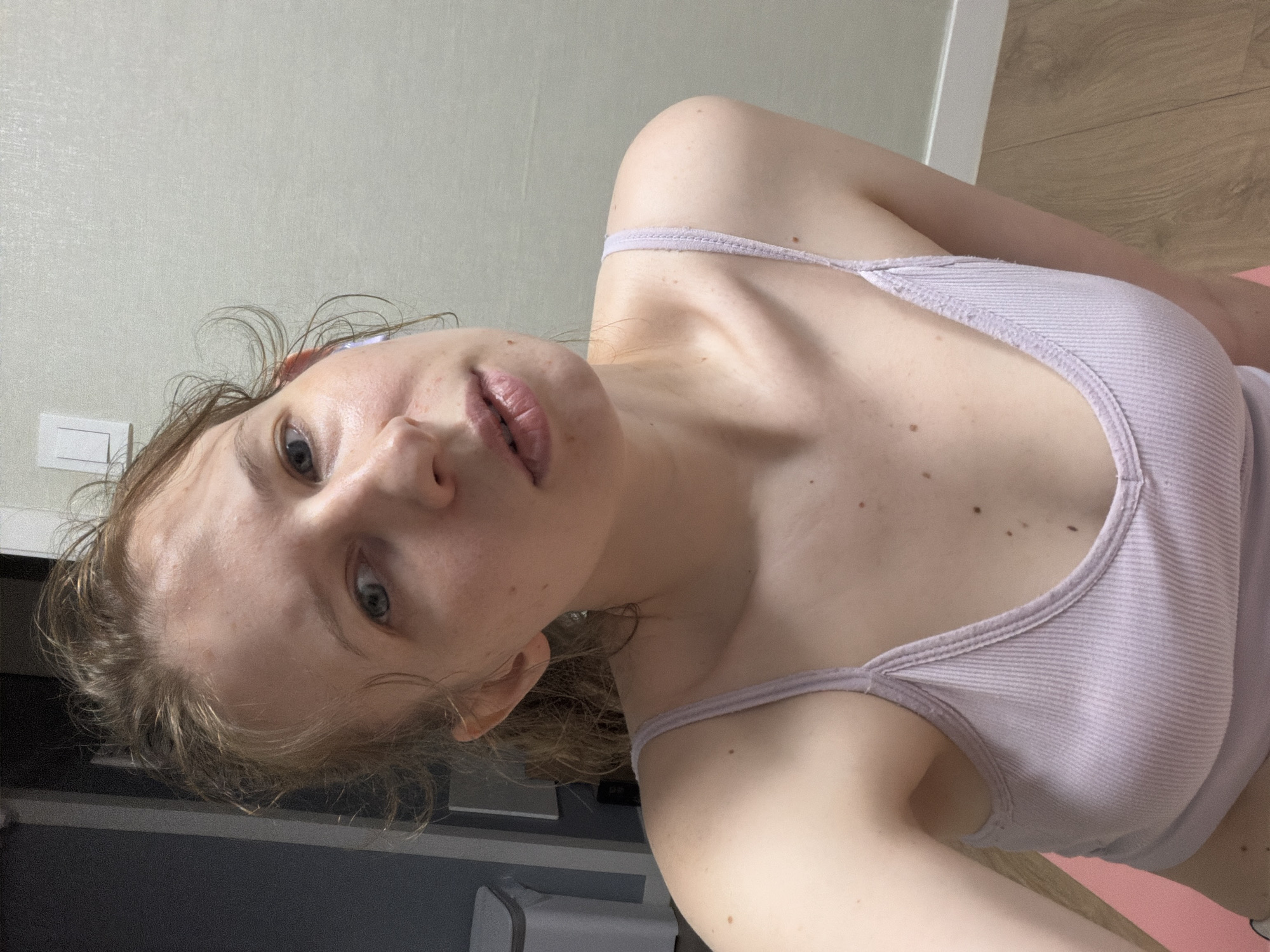 The image is a portrait of a young woman with fair skin, light brown hair, and blue eyes, taken from a low angle. She is wearing a light purple ribbed tank top. Her hair is slightly messy and falls around her face and shoulders. The background consists of a pale wall with a white electrical outlet on the left side, and a wooden floor with a pink mat on the right. The lighting suggests it is daytime. The woman's expression is neutral, and she appears to be looking directly at the camera. The setting appears to be indoors, possibly a home or a gym, given the presence of a yoga mat. There is no visible text in the image.