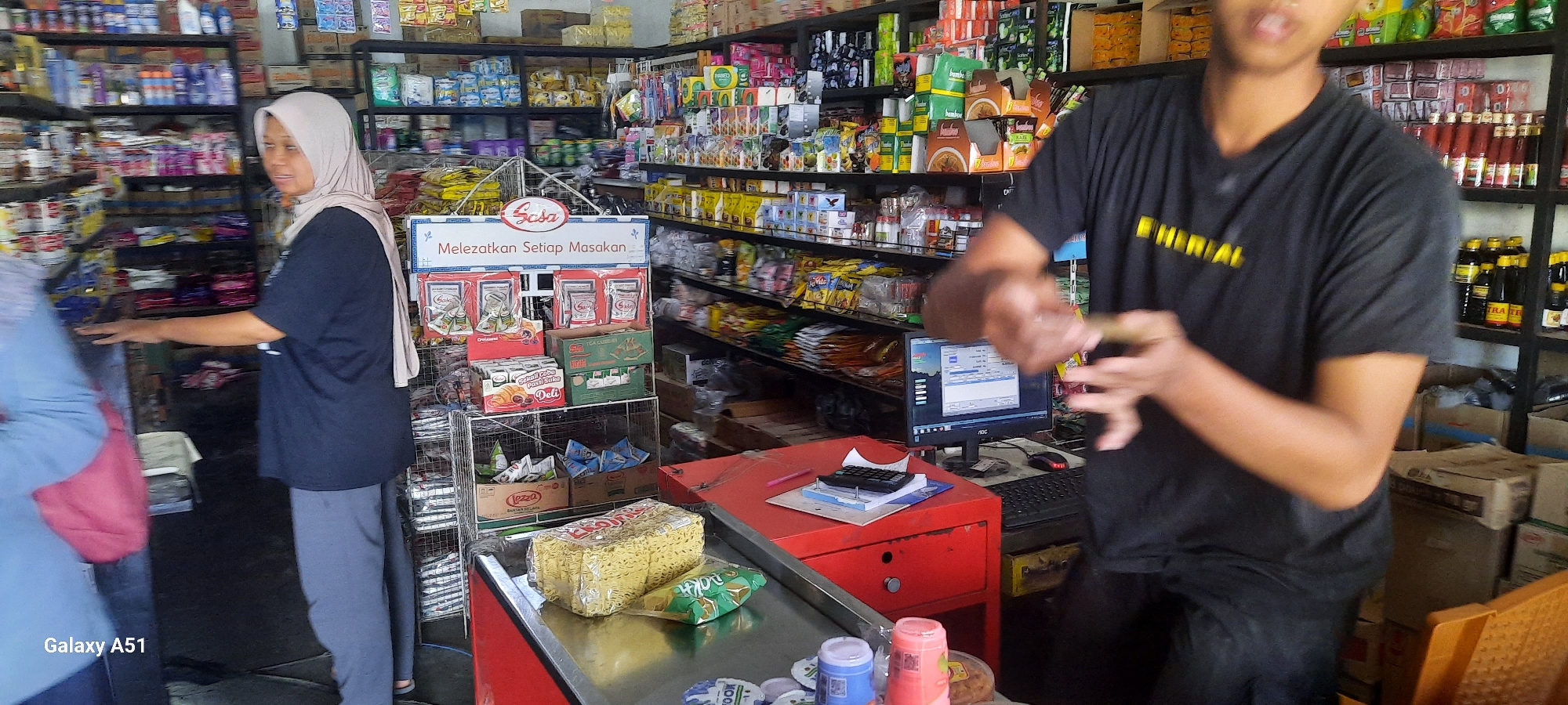 The image captures the interior of a small grocery store, likely a "warung" or minimarket, in Selopuro, Indonesia. The scene is bustling, with at least two individuals present. A woman wearing a light-colored hijab and a dark t-shirt is standing behind a counter, looking to her left. Her right hand is extended as if reaching for or displaying items. In the foreground, a person in a dark t-shirt with yellow lettering, possibly a cashier or employee, is partially visible, their hands in motion, suggesting an interaction such as handling money or scanning items. The store is densely stocked with a variety of products displayed on shelves lining the walls and on freestanding racks. Visible items include packaged food products, beverages in bottles, canned goods, and cleaning supplies. A red counter with a calculator and papers sits in the mid-ground, next to a computer monitor displaying a software interface. The overall impression is that of a typical Indonesian neighborhood store.