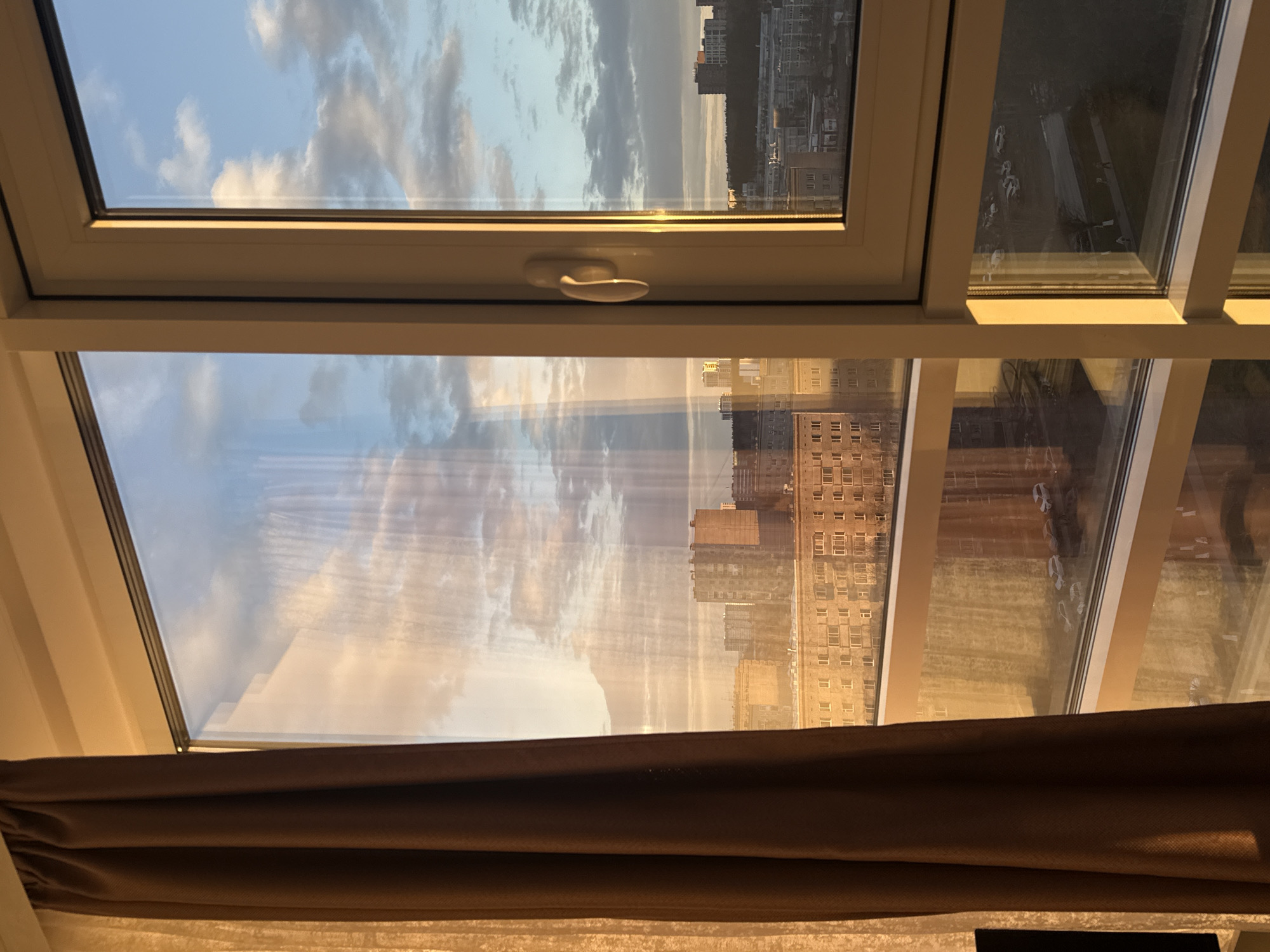 The image captures an indoor perspective looking out through a multi-pane window during the golden hour, likely sunrise or sunset. The window frame is white, and a white handle is visible on one of the vertical mullions separating the panes. Dark brown curtains are partially drawn at the bottom of the window.
Through the upper window sections, a clear blue sky with scattered white and grey clouds is visible, bathed in a warm, golden light. This same golden illumination reflects strongly on the glass panes and highlights the urban landscape outside.
The lower sections of the window present a view of a cityscape dominated by numerous multi-story buildings, some with a pattern of many small windows, suggesting older architectural styles. These buildings are also aglow with the soft, warm light. In the distance, a darker area, possibly a forested zone or further urban development, can be seen. Reflections of the radiant sky and clouds are prominent on the window glass, overlaying the view of the buildings. No people or visible text are present in the image.