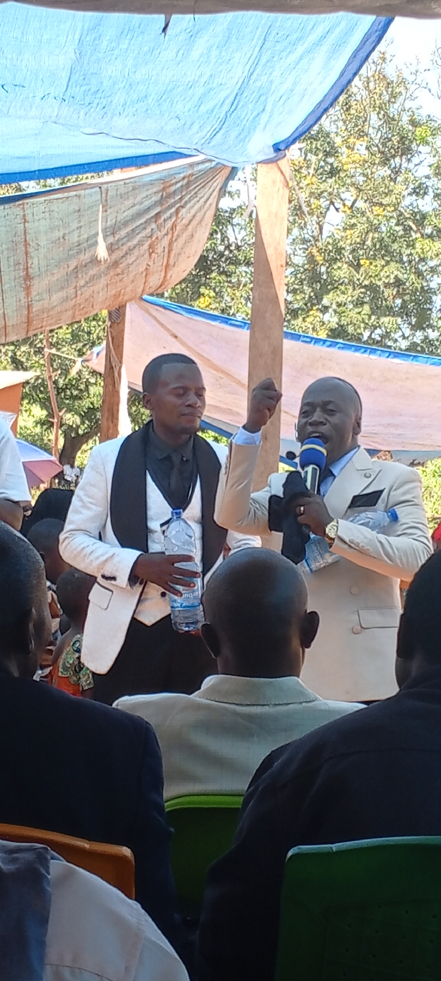 Outdoors in Kalemie, Democratic Republic of the Congo, two men in suits stand before a seated audience. The man on the left wears a white suit with black lapels and holds a water bottle. The man on the right, wearing a light-colored suit, speaks into a blue microphone and holds a black cloth and another water bottle. An overhead canopy provides shade. The audience consists of people in various attire, seated in green chairs. Trees are visible in the background.