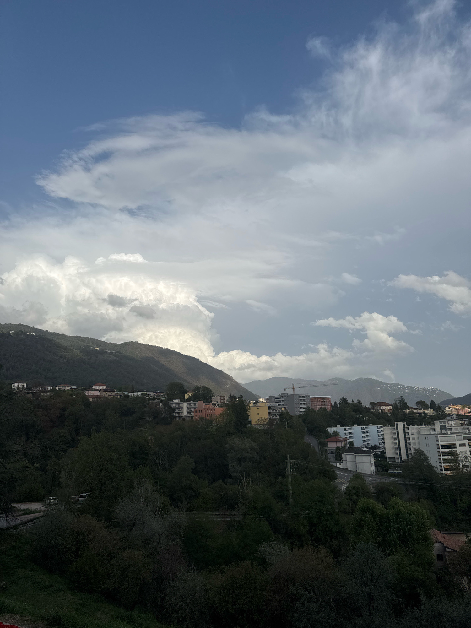 The image depicts a view of a town nestled in a valley with mountains in the background. The sky is a mix of blue and white clouds, with a large, dramatic cumulus cloud formation dominating the upper left portion of the frame. In the mid-ground, several multi-story buildings are visible, some with light-colored facades, others in shades of pink and yellow. A construction crane is present near one of the buildings. The foreground is filled with dense, dark green trees and foliage. A winding road with some vehicles is visible amongst the trees in the lower left section of the image. The overall scene suggests a cloudy or partly cloudy day in a hilly or mountainous urban environment.