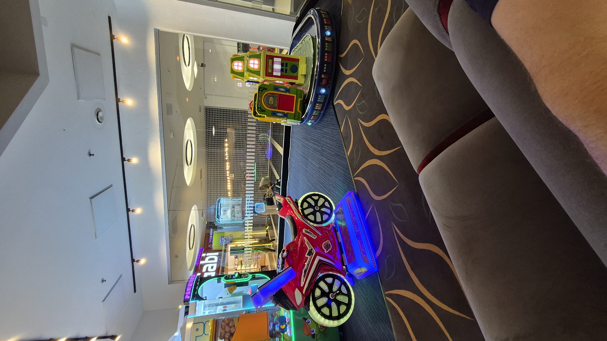 The image captures an indoor amusement area, likely a section of a larger establishment such as a mall or entertainment center, with no specific indicators of it being in Daugavpils, Latvia. In the foreground, two plush, grey couches are visible, partially obscuring the view of the area beyond. The floor is covered with a dark carpet featuring a repeating pattern of stylized yellow leaves.
The focal point of the scene is an array of children's amusement rides. To the left, a bright red, motorized motorcycle ride for children is prominently displayed. It features illuminated wheels with colorful glowing rims and a blue platform at its base, also lit with blue lights. Behind the motorcycle, a claw machine or similar arcade game with a yellow and clear casing is partially visible.
To the right of the motorcycle, a miniature train-themed ride is set up on a circular track. The ride consists of several colorful, box-like carriages, designed to resemble a train, with a small, two-story structure resembling a house on top. The track itself is bordered by a strip of colorful train car graphics.
The background of the image reveals a brightly lit interior with large glass windows or partitions. Through these, more arcade games and an interior space with shelves and displays, possibly a shop, can be seen. The ceiling features modern track lighting with multiple bulbs and recessed circular light fixtures. The overall impression is that of a clean, well-lit, and engaging children's entertainment space.