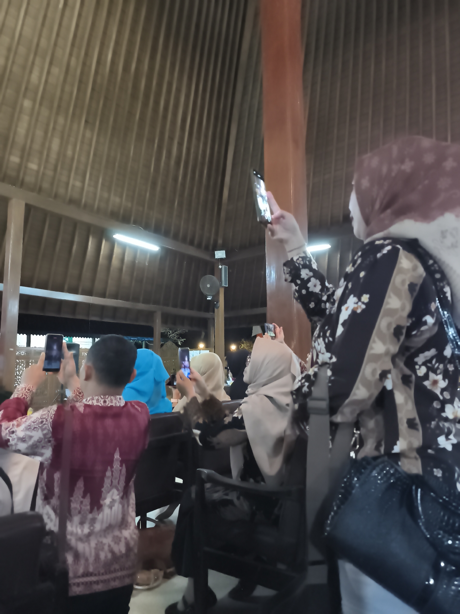 The image depicts a group of people, possibly attending an event, indoors with a distinct wooden ceiling structure. Several individuals are holding up smartphones, suggesting they are taking pictures or videos. One person in the foreground, a woman wearing a headscarf and a patterned dress, is seen holding a phone with her right hand. To the left, a young boy in a red and white patterned shirt is also holding a phone. Other people, also wearing headscarves, are visible in the background. The environment appears to be a communal space, possibly a hall or a restaurant, with wooden pillars and ambient lighting. The overall atmosphere is one of engagement with an ongoing activity, captured through personal devices. The location is identified as Banyumas, Indonesia.