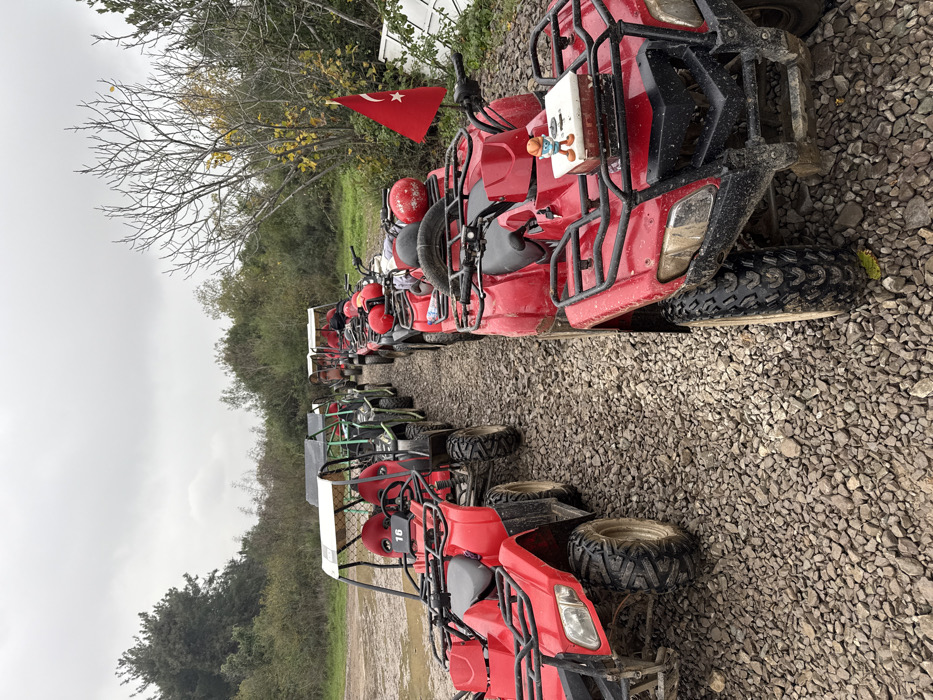 The scene is an outdoor staging area for off-road vehicles in Çiftlikköy, Turkey, under an overcast sky. A gravel path serves as the parking surface for multiple ATVs and UTVs/buggies, arranged in two irregular lines. The majority of the vehicles are red, with a few green UTVs visible in the background of the left line.
In the foreground, a red ATV on the right displays a small Turkish flag attached to its handlebar and a small, orange-capped figurine standing on a white box secured to its front cargo rack. Several red helmets are placed on the seats or cargo racks of the ATVs in this line, indicating preparation for or completion of a recreational activity. Another red ATV in the left foreground, behind the first, exhibits the number "16" on its front panel. All vehicles show evidence of prior use, with mud and dirt adhering to their tires and lower chassis.
The environment comprises dense, varied foliage and trees, some with bare branches or yellowing leaves, lining the sides of the gravel path. In the far right background, partially obscured by the trees, some discarded white panels or objects are visible on the ground. No individuals are present in the image.