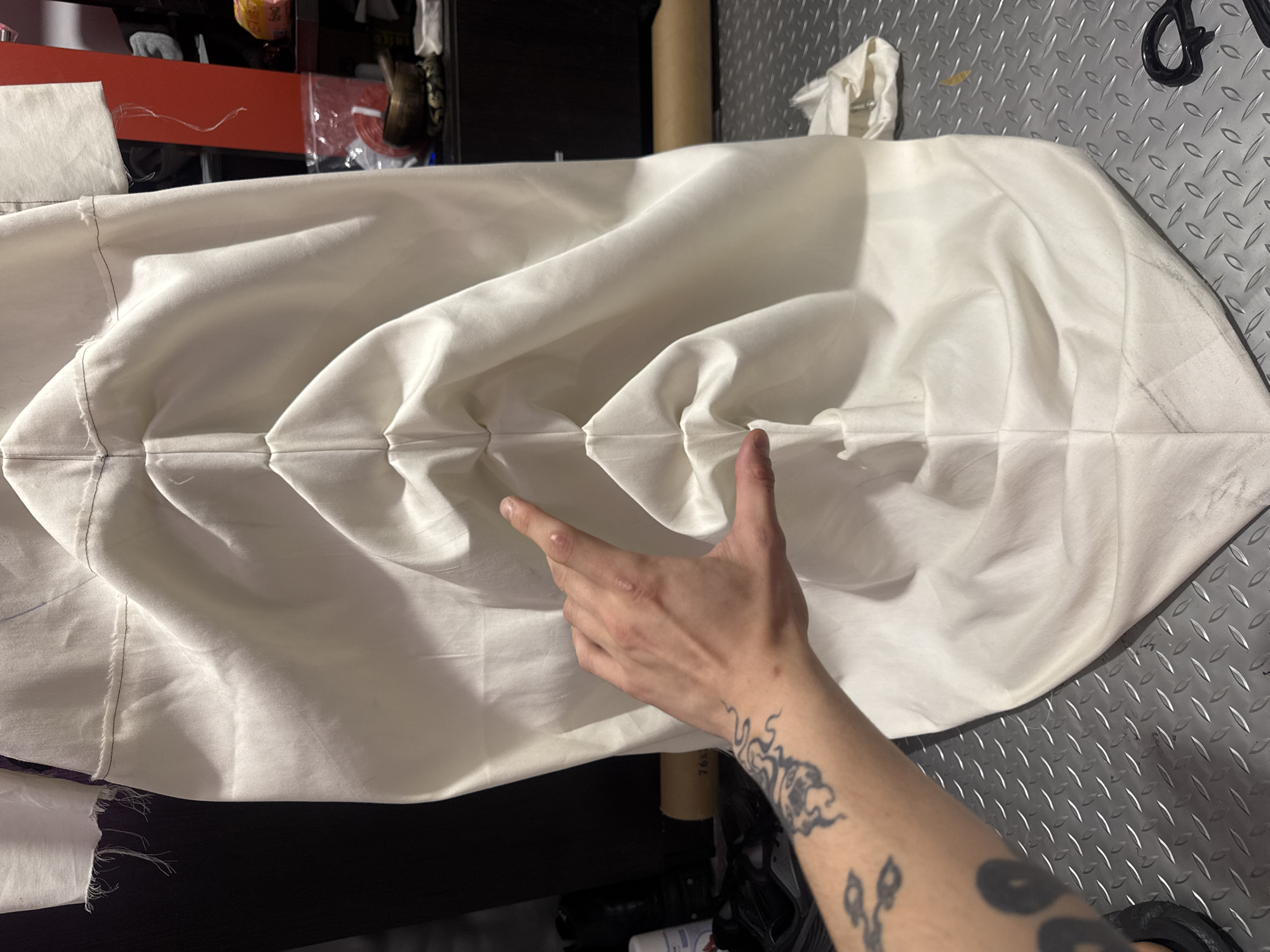 The image displays a close-up view of a person's arm and hand interacting with a piece of white fabric, likely within a workshop or creative studio in Sofia, Bulgaria. The main subject is a section of white textile, characterized by prominent horizontal pleats or folds across its width, creating a textured, undulating surface. Visible stitching, some frayed, runs along the seams of the fabric, and faint pencil or chalk markings are discernible on its surface, indicating it is a work in progress.
A person's right arm, adorned with extensive dark tattoos, extends into the frame from the lower right, with their hand gently touching or gesturing towards a fold in the fabric. A small scar or mark is visible on the knuckles of the index finger.
The background shows elements typical of a workspace: a partially visible red shelf, various indistinct objects, and a roll of brown material with the text "76X" printed on it. The surface beneath the fabric is a metallic diamond plate pattern, and a pair of black scissors can be seen on this surface in the upper right. The overall setting suggests a practical environment for textile work.