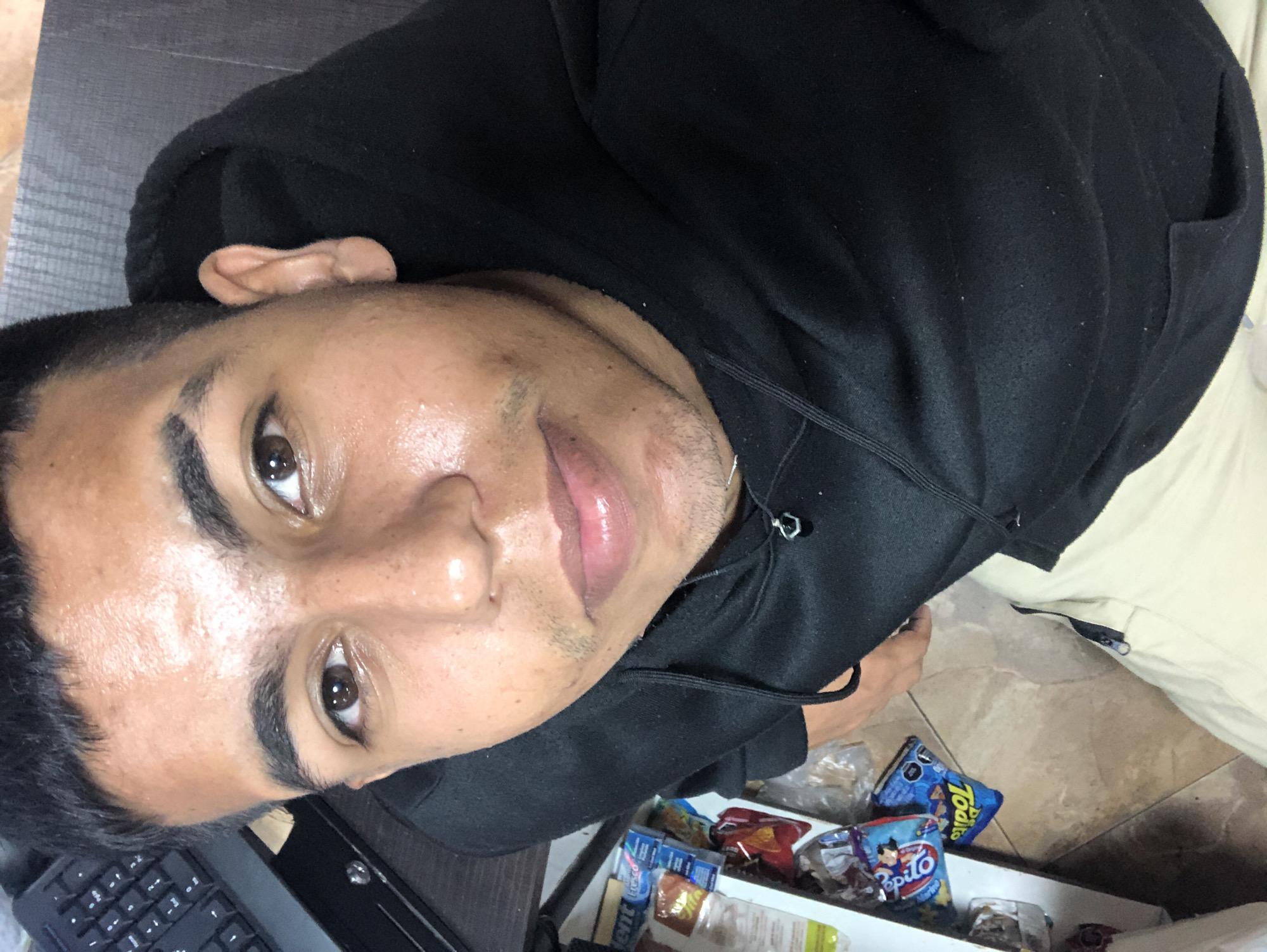 A close-up, slightly rotated portrait depicts a young man wearing a black hooded sweatshirt. He has dark, short hair, dark eyes, and tan skin. A portion of a black keyboard is visible on the left side of the frame. Below the man, there is a shelf containing various snack items, including packages of "Toditos" and "Pepito" chips. Light-colored pants are visible on the right side. The location is not identifiable as being in Juan Griego, Venezuela, from the visible elements in the image.