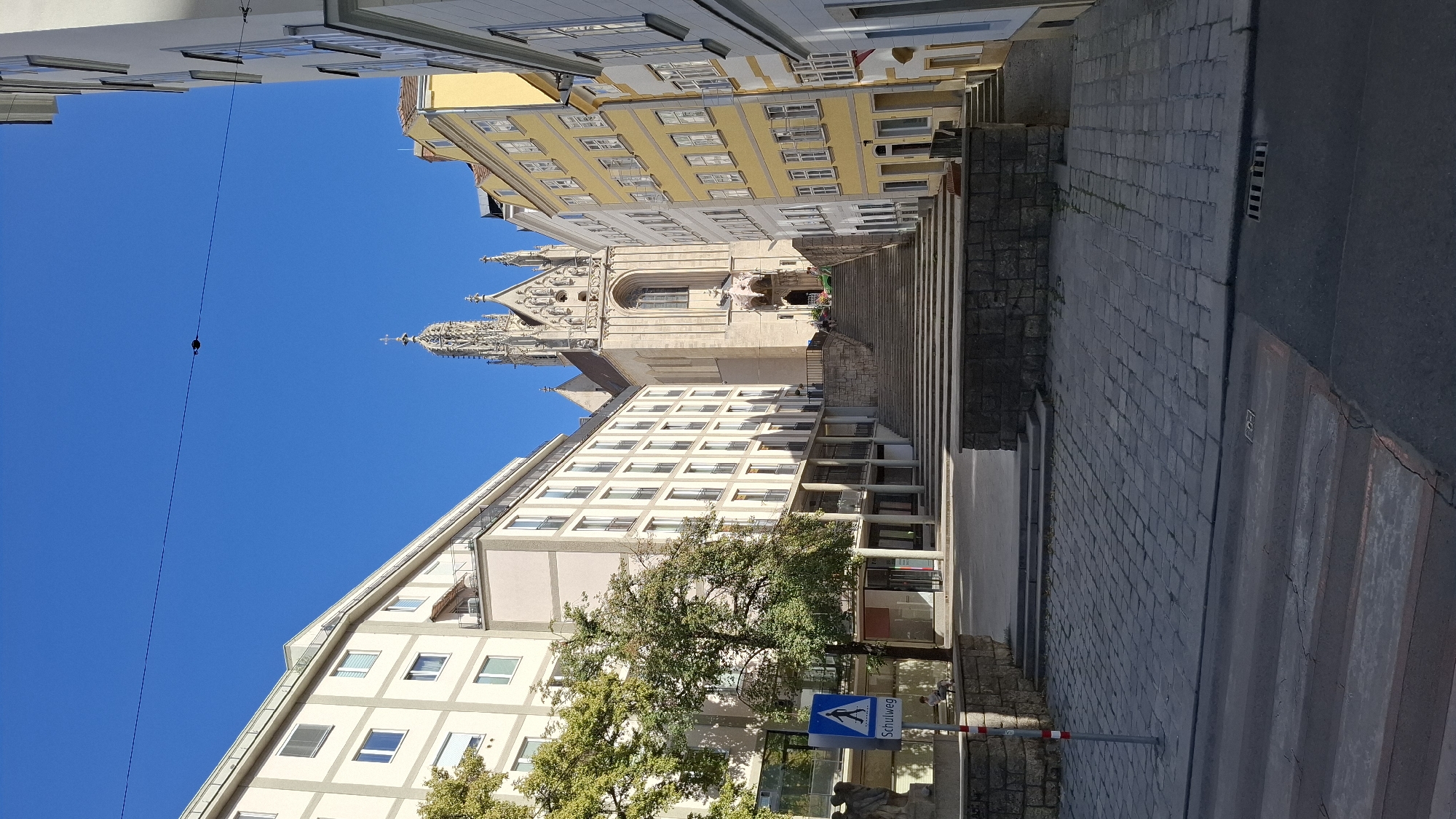 The image depicts the Peterskirche (St. Peter's Church) in Vienna, Austria, viewed from a street level perspective. The church, with its distinctive spire and ornate facade, is situated at the top of a wide, stone staircase that dominates the center of the frame. The staircase is flanked by modern buildings on either side. The building on the left is a multi-story structure with numerous windows, appearing to be residential or office space. The building on the right is painted yellow and also has many windows, with traditional European architectural features.
In the foreground, a cobblestone street is visible, with a pedestrian crossing marked by white stripes. To the left of the staircase, a blue sign with a white pedestrian symbol and the word "Schulweg" (school path) is mounted on a pole. A large tree with green foliage stands behind the sign. The sky is clear and blue.
Several people are visible in the distance, ascending the stairs towards the church entrance. The overall scene suggests a sunny day in a European city, with a mix of historical and modern architecture. The perspective is from a street level, looking uphill towards the church.