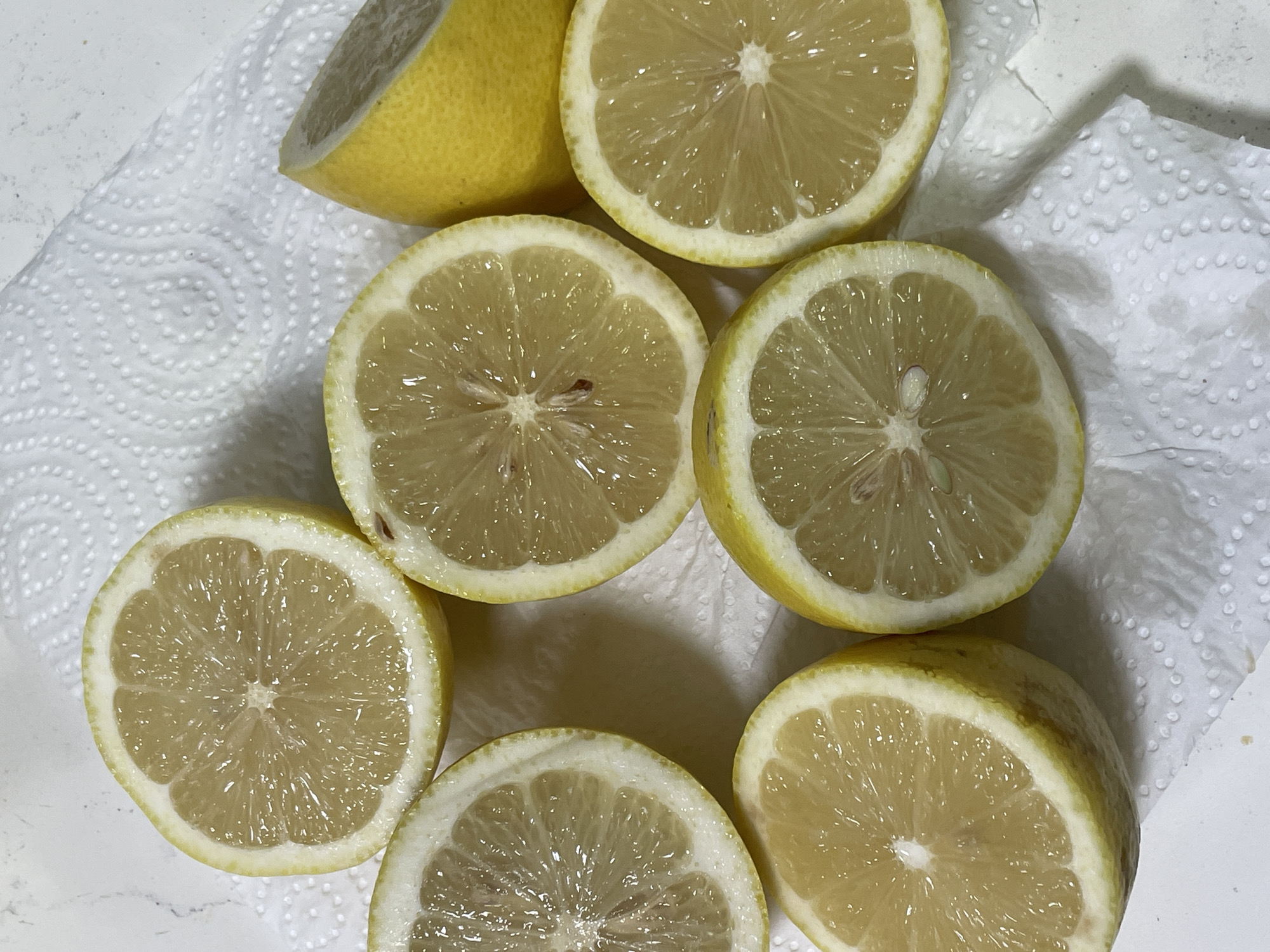 Six lemon halves and one whole lemon piece are arranged on a white paper towel. The lemons are cut crosswise, revealing their juicy interiors and seeds. The scene is indoors, on a white surface that appears to be a countertop. The lighting suggests an overhead light source. The image provides no information about the city of Raanana, Israel.