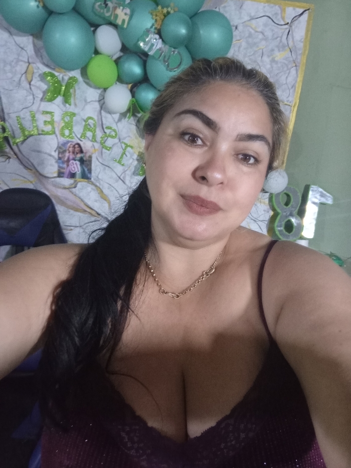 A middle-aged woman with light skin and dark hair pulled to one side poses for a selfie. She wears a low-cut, dark top and a gold necklace. Behind her is a decorated wall with green and white balloons, green lettering spelling "Isabella", a small photo, and a silver number "81". The background suggests a celebratory event.