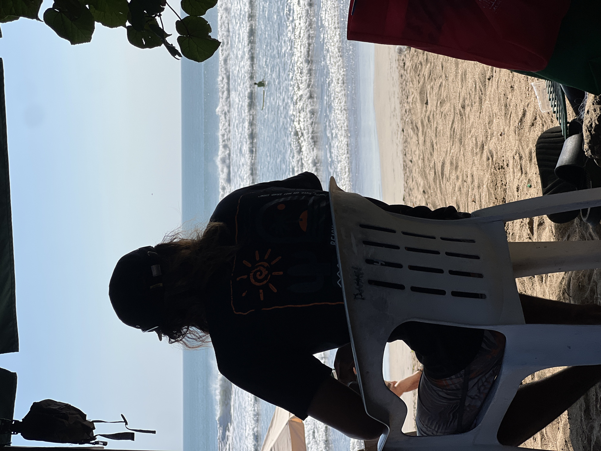 A person wearing a black t-shirt, shorts, and a baseball cap is seated in a white plastic chair, facing the ocean. The person's back is to the camera, and they appear to be looking out at the water. The ocean has moderate waves, and a surfer can be seen in the distance. The scene is on a sandy beach, with a beach umbrella visible to the left. The time of day appears to be late afternoon, with the sun casting long shadows. The location is Kuta, Indonesia, a popular tourist destination known for its beaches and surf. The overall mood of the image is peaceful and relaxed.