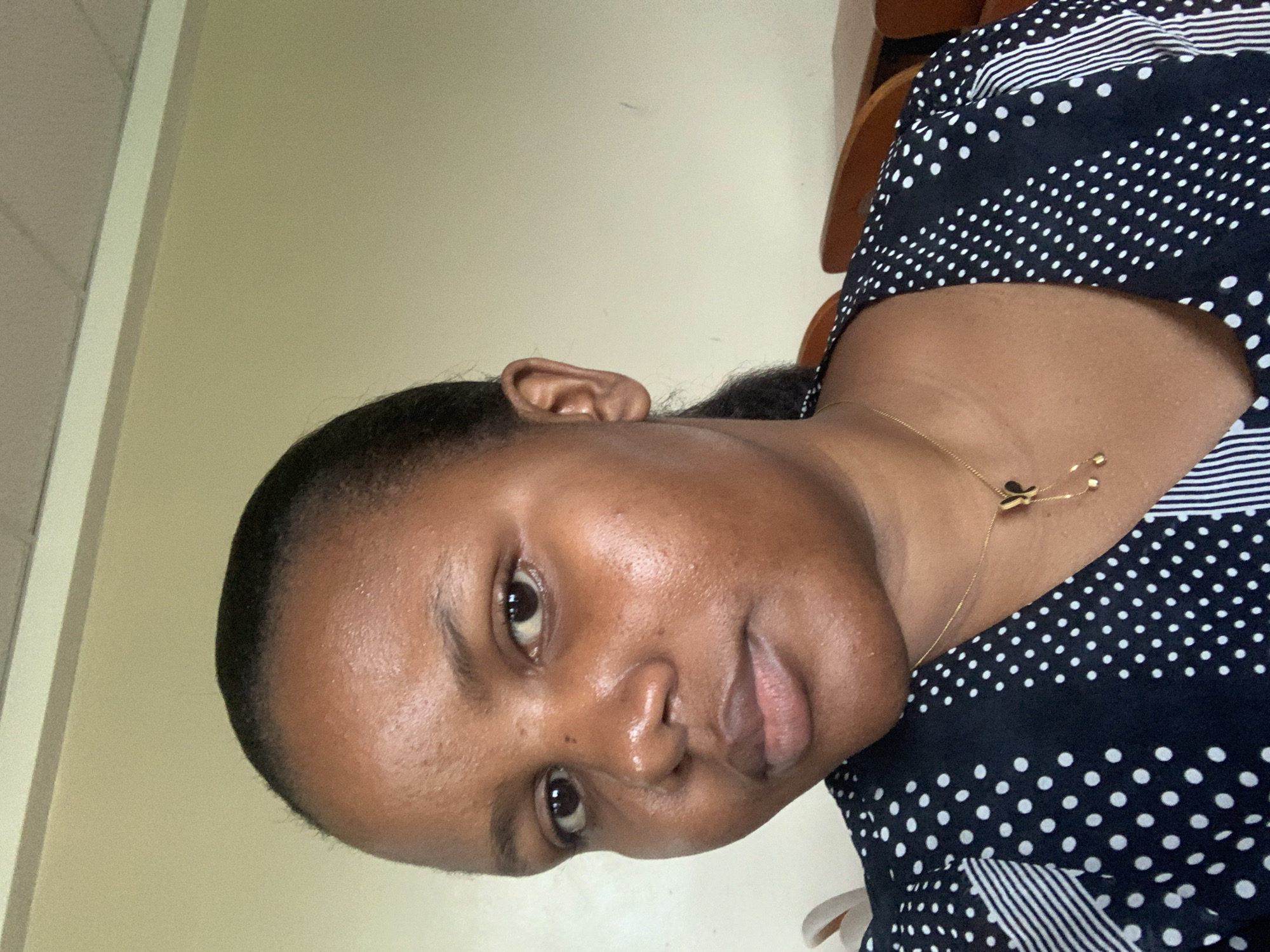 Here is a detailed description of the image:
**People:** A dark-skinned woman is the primary subject.
**Objects:** She's wearing a dark blue and white polka-dot blouse and a thin gold necklace with butterfly charms.
**Environment:** The background appears to be an indoor setting with a light beige wall. Some wooden furniture is visible in the periphery.
**Location:** It is not possible to determine if this image was taken in Dar es-Salaam, Tanzania. No information related to location is present in this image.