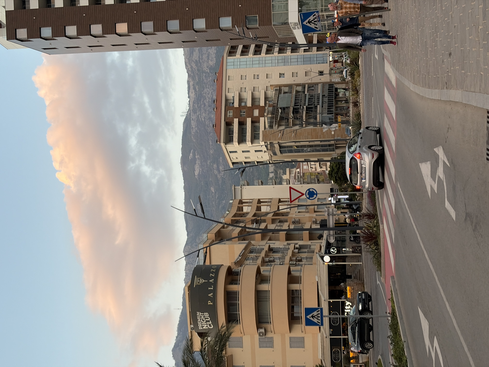 The image depicts a street scene in Budva, Montenegro, with a building identified as "Palazzo Premium Night Club" visible in the foreground. A mountain range serves as a backdrop. A silver car is positioned on the right side of the street, and a crosswalk is visible. Pedestrians are crossing the street. Another car is parked near a restaurant. Street signs and lamp posts are present, indicating an urban environment. The sky is partly cloudy with a sunset cast.