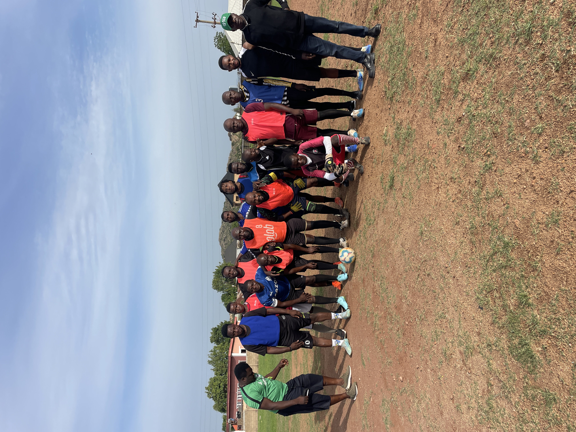 A medium shot depicts a group of approximately 17 men posing outdoors on a dirt and grassy area, presumed to be a field. The sky is blue with some clouds. A few men in the front are posing with soccer balls. Most men are wearing athletic jerseys. One man at the far right is in a black jacket. To the left, one man is wearing a green shirt. Trees and buildings are visible in the background. Utility poles are visible. The image is likely taken in Jos, Nigeria.