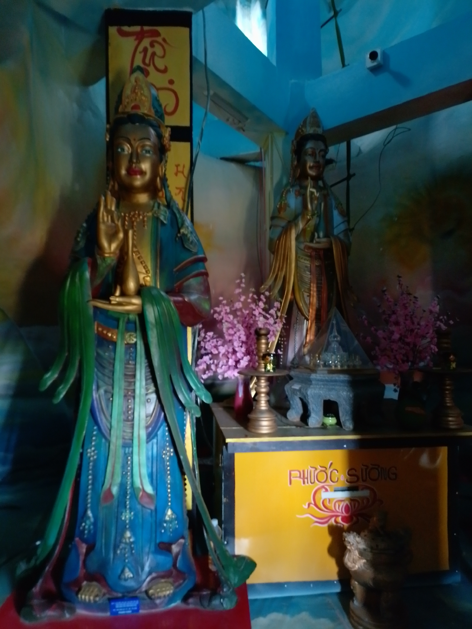 The image shows two statues in an indoor setting in Unknown, Unknown. The statue on the left is taller and more ornate, with a gold-colored face and hands, and wearing a blue and green robe with intricate details. It holds a small gold vessel in its left hand and raises its right hand in a gesture. Behind it is a yellow panel with red and black lettering in an unknown language. The statue on the right is smaller, with a similar gold-colored face and hands, and wearing a light blue and gold robe. It also holds a small object in its hands. Between the statues are small pink flowering plants in pots, and two golden candle holders. The statue on the right sits atop a gray stand, which is on a yellow platform with red and white lettering and a decorative design. The walls and ceiling are a light blue color, and there is a security camera visible in the upper right corner.
