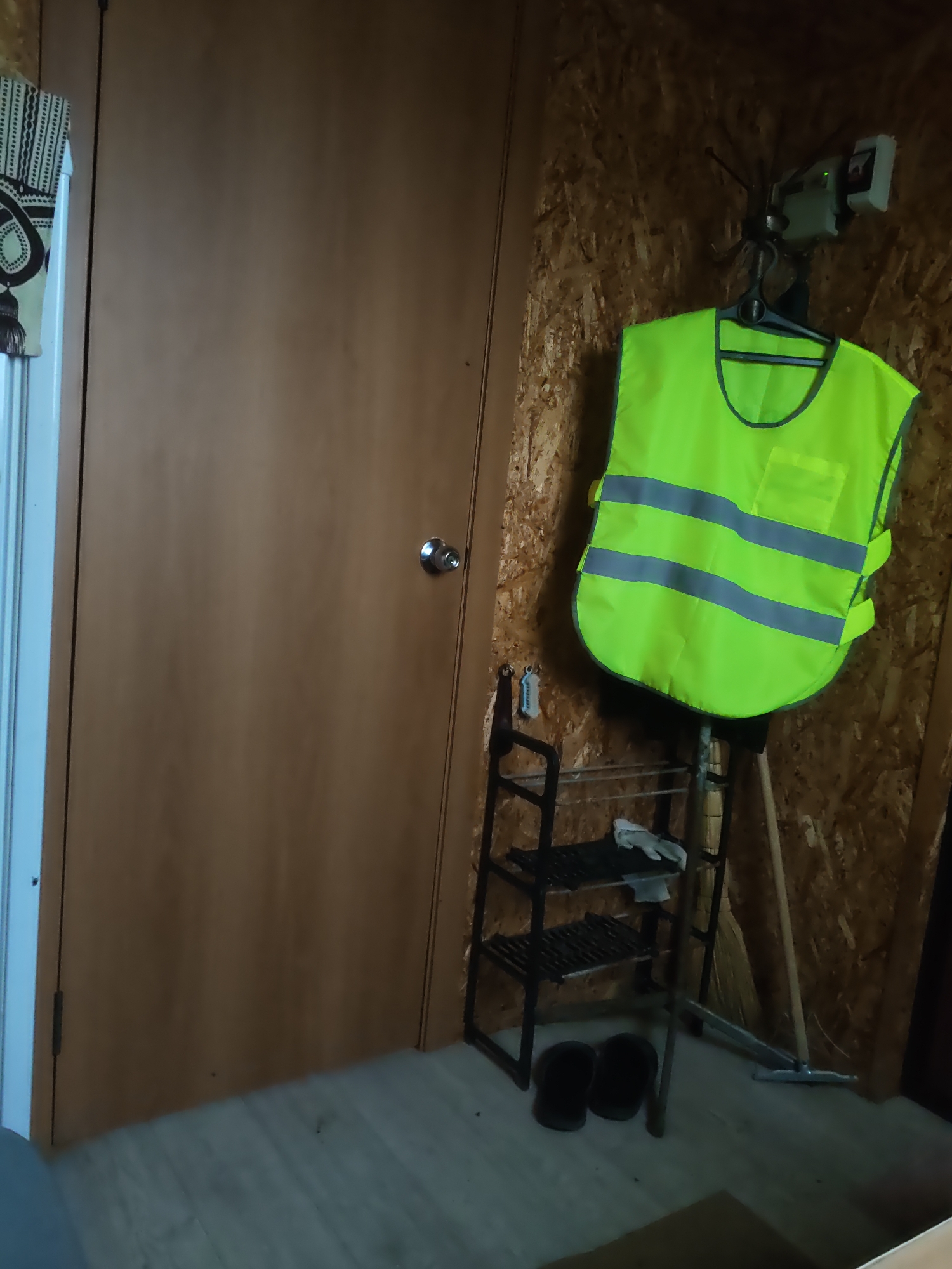 The image depicts a small room, possibly a storage area or workshop, with a light brown wooden door on the left side. The door has a silver round doorknob. The walls of the room are made of textured particleboard. On the right side of the room, a bright neon yellow safety vest with gray reflective stripes is hanging on a hook. Below the vest, there is a black metal shelving unit with two shelves, holding a pair of white work gloves on the top shelf. Two black flip-flops are placed on the floor in front of the shelving unit. A broom and a shovel are leaning against the wall behind the shelving unit. The floor appears to be made of light-colored wooden planks. There is a curtain with a patterned design hanging on the left side of the image, partially obscuring the doorway. The lighting in the room is dim. The scene is consistent with a utilitarian space in a residential or small commercial building.