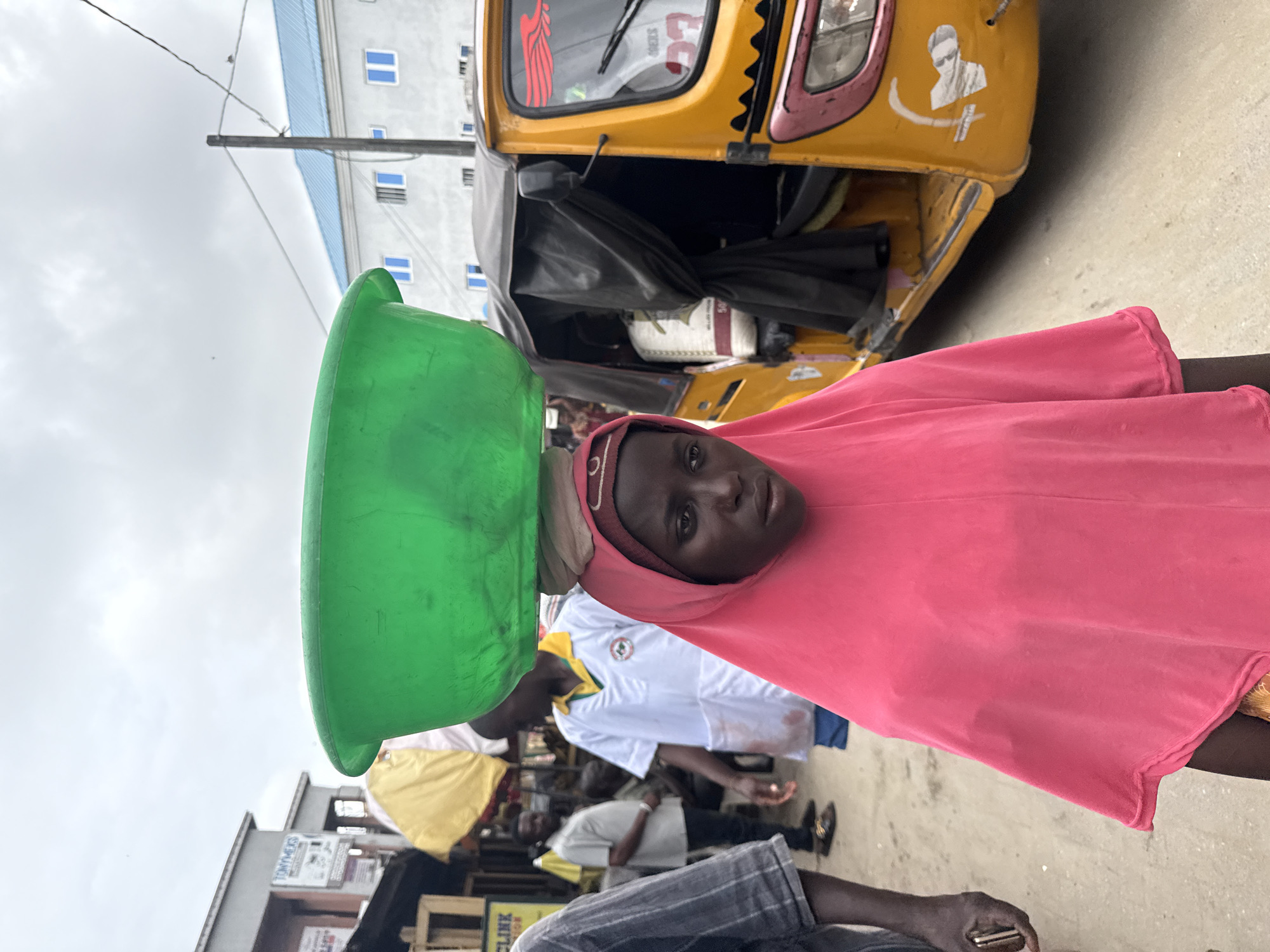 A young girl wearing a bright pink headscarf and covering her body with a pink hijab walks forward carrying a large green basin on her head. She is in a street that appears to be in Lekki, Nigeria. Behind her, a yellow auto-rickshaw is parked. The auto-rickshaw has black tarpaulin covering the rear compartment and the front of the vehicle is adorned with stickers. Other people are visible in the background, including a man in a white lab coat with a green collar. The ground is paved with light-colored dirt or sand. The sky is overcast.