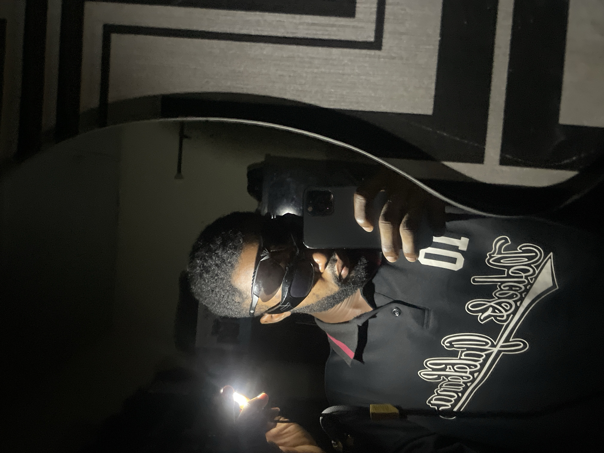 This image depicts a mirror selfie of a dark-skinned adult male in an indoor setting in Lokoja, Nigeria, under extremely low ambient light conditions. The subject is positioned to the left of a large, dark-framed rectangular mirror, partially visible on the right.
The man has short, dark, curly hair and a neatly trimmed beard. He is wearing dark, wrap-around sunglasses that obscure his eyes. His attire consists of a black polo shirt with a visible dark red or maroon trim on the inner collar edge. A stylized white logo, appearing mirrored, is printed on the left chest area, featuring script that appears to read "California" or similar, below which is the mirrored number "01".
In his right hand, the man holds a silver-colored smartphone with a visible triple-lens camera array, positioned as if taking the photograph. In his left hand, raised slightly above his head, he holds a small, rectangular LED flashlight emitting a bright, concentrated white light that serves as the primary illumination source for the scene.
The background reflected in the mirror, beyond the subject, is largely dark on the left, while the right side reveals a wall with a prominent geometric pattern composed of dark, thick lines forming rectangular shapes against a lighter gray surface. The overall environment is dim, relying heavily on the direct light from the flashlight, suggesting a power outage or intentional low-light scenario within the Lokoja location.