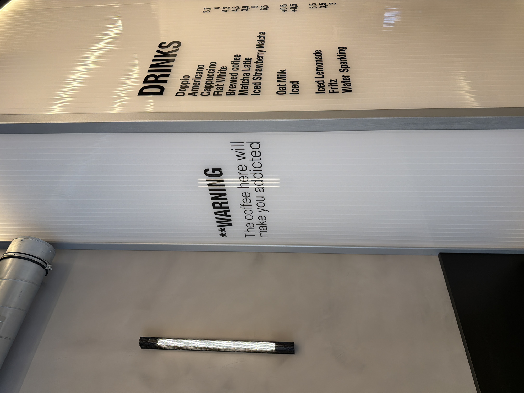The image captures a detail of a coffee shop menu, which includes a warning: "**WARNING The coffee here will make you addicted." Above this, the menu lists various drinks and their prices, including Doppio (3,7), Americano (4), Cappuccino (4,2), Flat White (4,8), Brewed coffee (3,9), Matcha Latte (5), Iced Strawberry Matcha (6,5), Oat Milk (+0,5), Iced (+0,5), Iced Lemonade (5,5), Fritz (3,5), and Water Sparkling (3). The menu is mounted on a wall with a horizontal light fixture. There's also a visible pipe or ventilation system in the scene. The overall setting suggests an interior space, possibly a café or coffee shop in Wiesbaden, Germany.