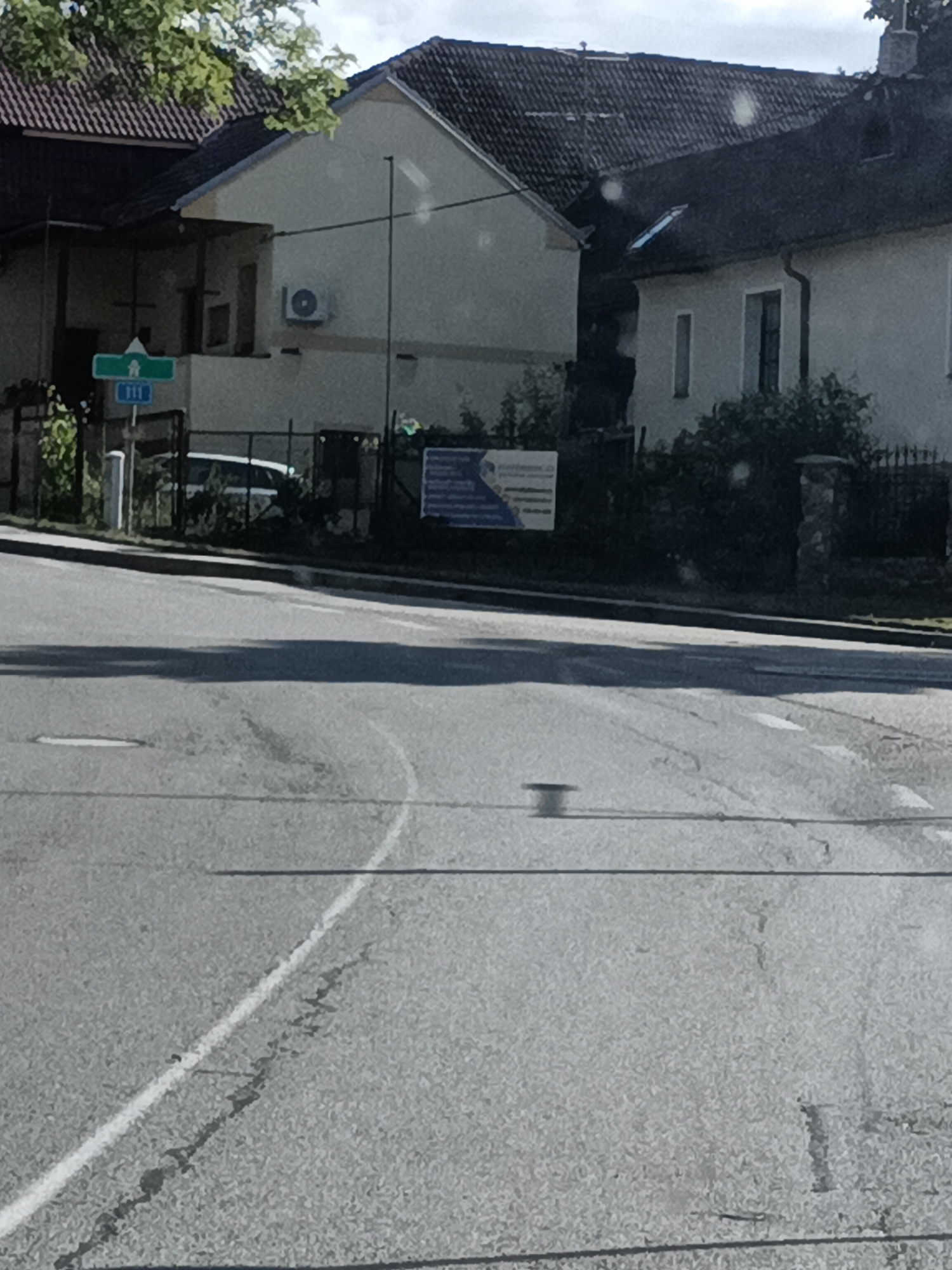The image shows a street in the city of Dalovy, Czechia. A white car is parked behind a fence. A street sign with the number 311 is visible. There are houses and trees in the background. The sky is cloudy. The image appears to be taken from a moving vehicle, as indicated by the blurriness of the road.