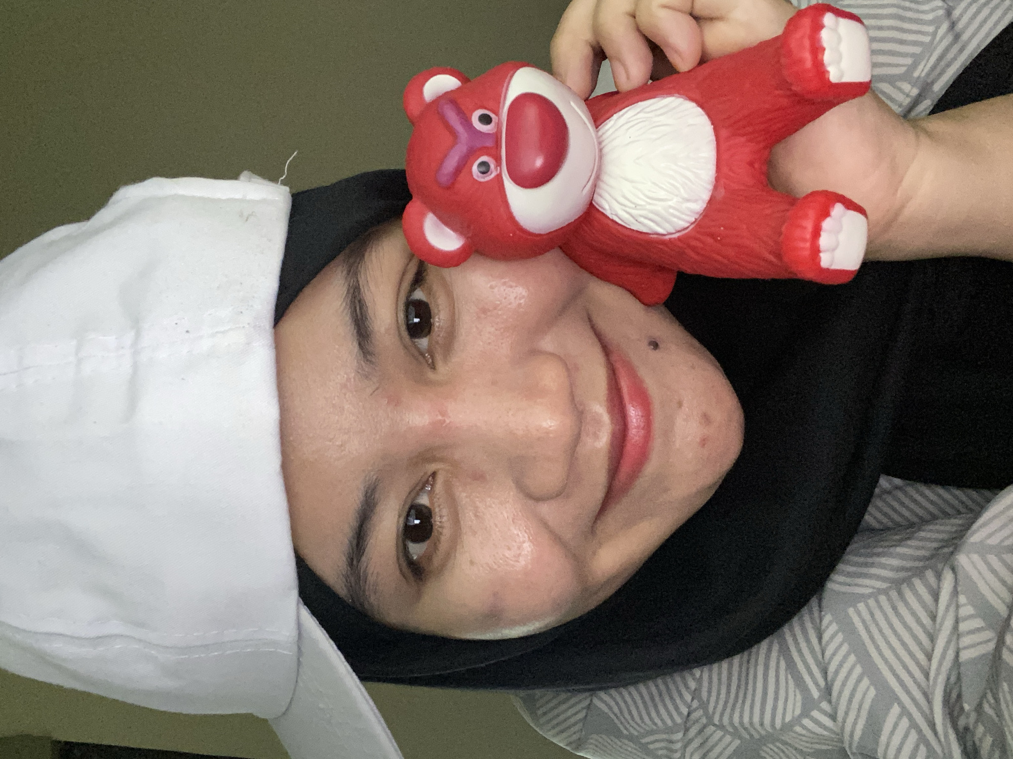 A medium shot captures a young woman in a white baseball cap and a black hijab, holding a red toy bear with angry eyebrows. She is looking towards the camera with a slight smile. The background is a plain, dark wall. The image is cropped from the shoulders up.