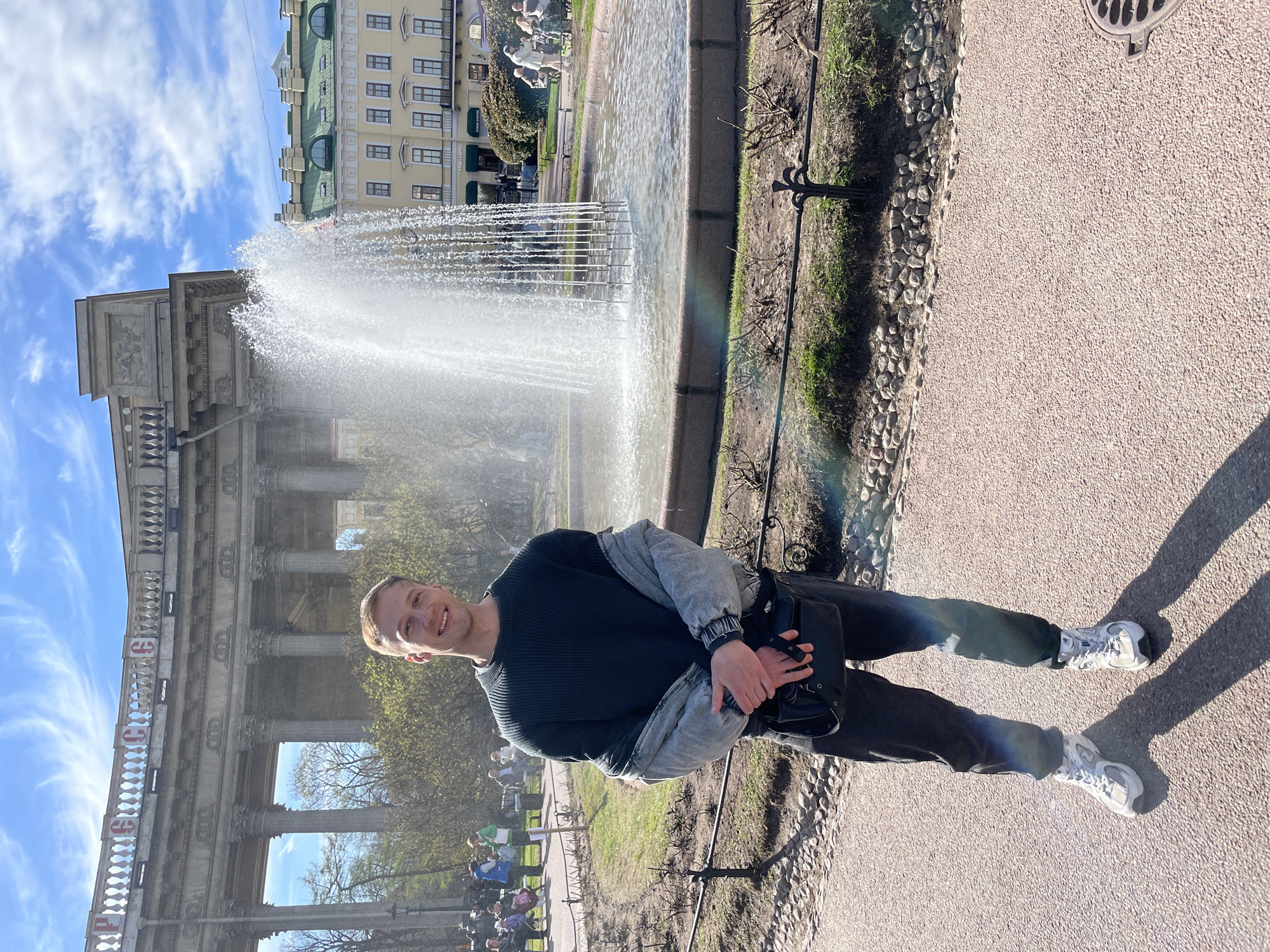 A man stands next to a fountain in Saint Petersburg, Russia. He wears a dark sweater, gray jacket, dark pants, and white sneakers. He is holding a black bag in his hands. Behind him is a large fountain with water spraying upwards. To the left of the fountain is a grand building with columns and architectural details. In the background, a lighter colored building and blue sky with clouds are visible. The man is standing on a paved path next to a grassy area.