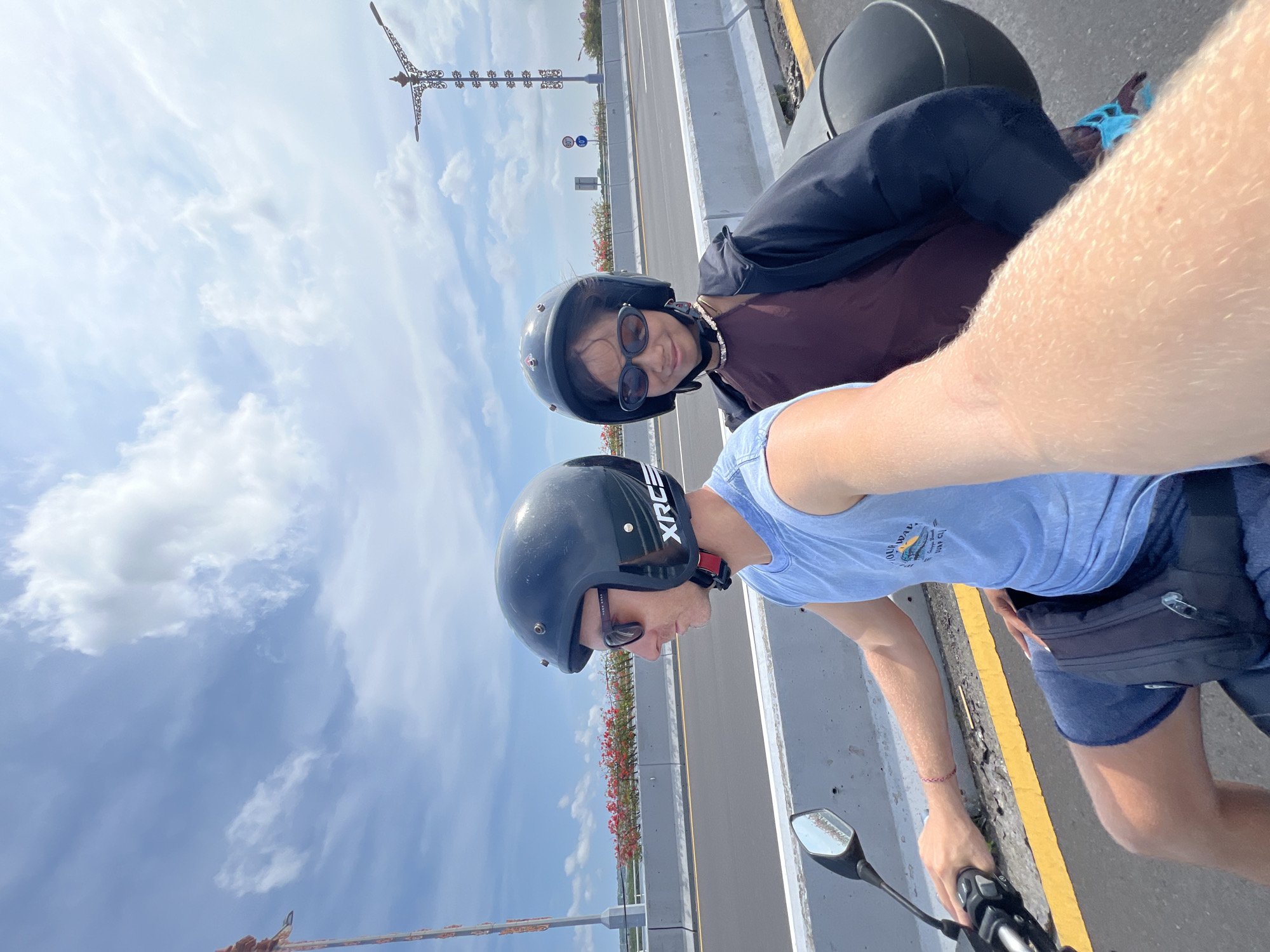 Two people wearing helmets are riding a motorcycle on a bridge in Nusa Dua, Indonesia. The foreground includes the rider's arm, hand on the motorcycle handle, and side mirror. Both are wearing helmets. The rider is in a blue t-shirt and shorts. The passenger is wearing a brown top and dark jacket. A tall, decorative metal structure is visible in the distance. The sky is blue with scattered clouds. A yellow road line and flower-lined barrier separate the motorcycle lane from the road.