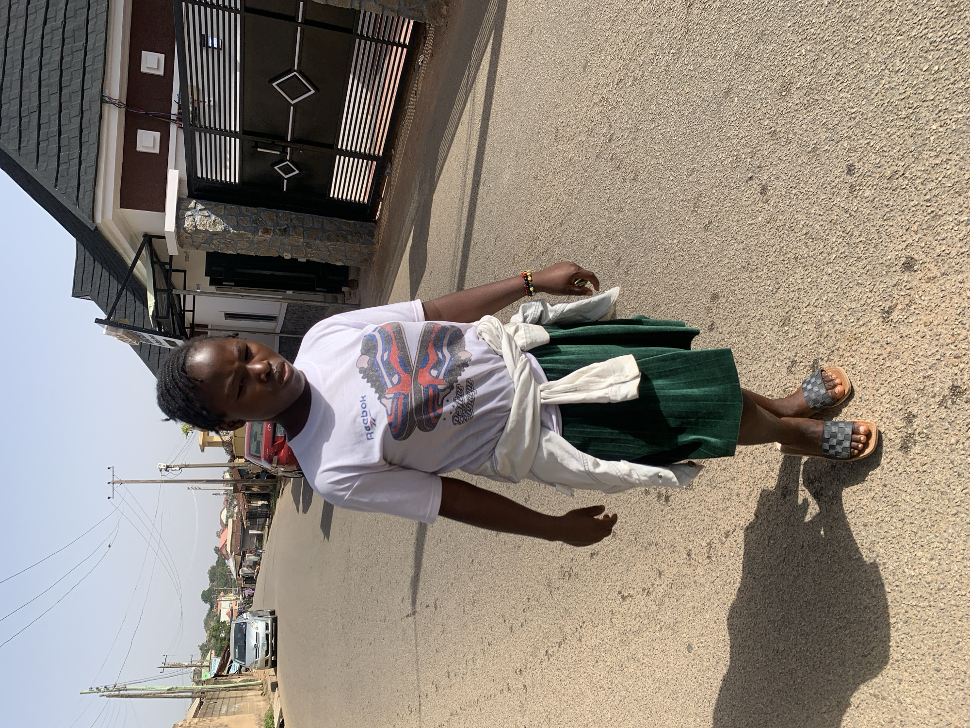 A woman stands on a paved road in Jos, Nigeria, based on the visible power lines and building styles. She wears a white t-shirt with a graphic print, a green skirt, and slides. A white jacket is tied around her waist. Behind her is a house with a dark-tiled roof, brown siding, and a black metal gate. In the background, power lines extend into the distance, and other buildings and vehicles are visible along the road. The lighting suggests it's daytime.