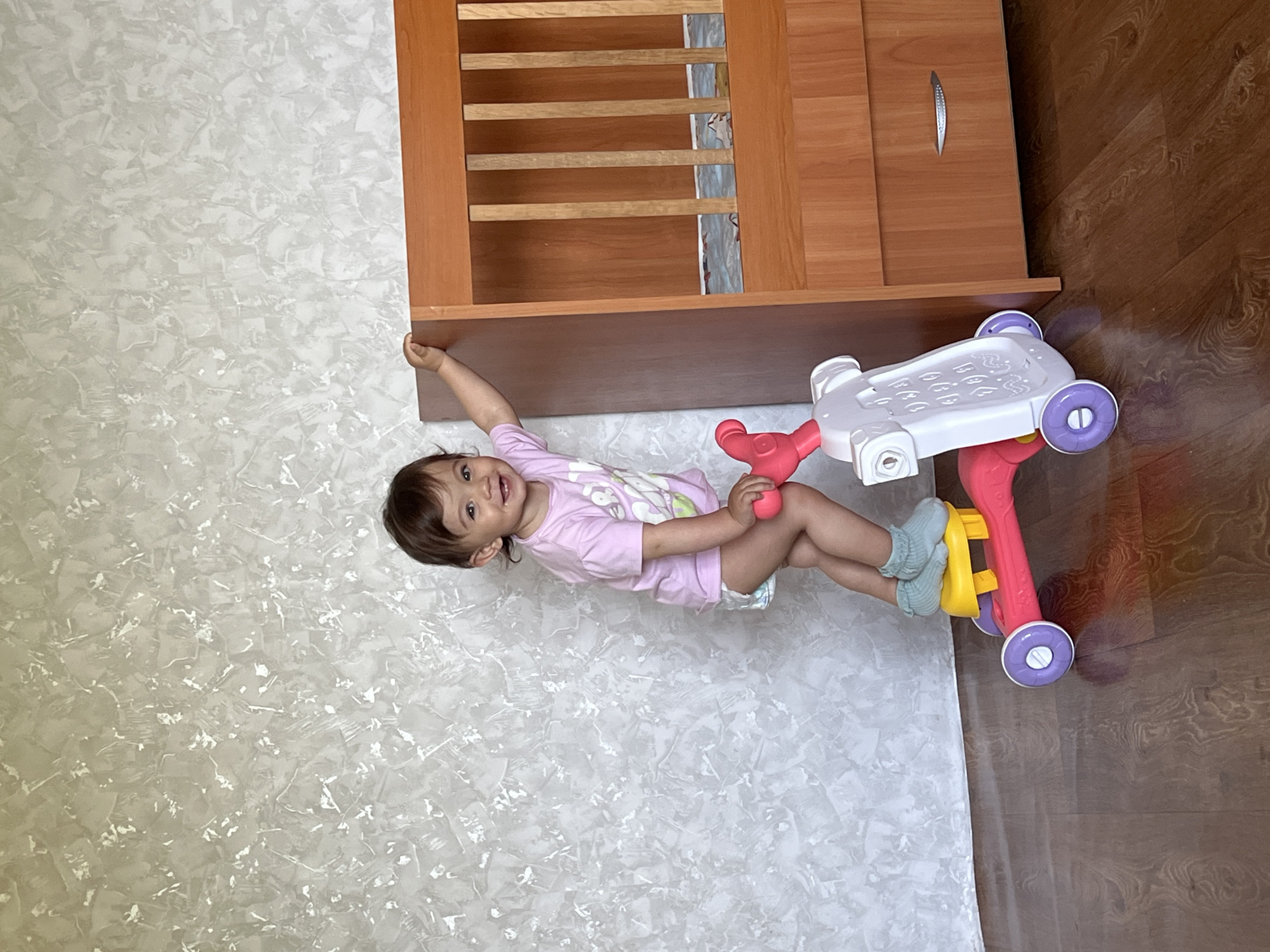 A young child with dark hair and a pink t-shirt stands on a pink and yellow toy scooter next to a wooden piece of furniture with a drawer and slats. The child is smiling and looking at the camera, with their right hand resting on the furniture. The background is a textured light gray wall and a dark wood floor. The scene appears to be indoors, possibly in a residential setting in Elena, Bulgaria, although the specific location is not identifiable from the image.