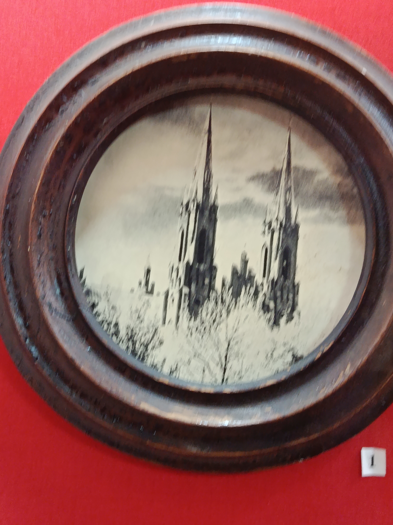 An oval, sepia-toned photograph in a dark brown, carved wooden frame depicts two Gothic-style church spires rising above a skeletal forest, likely photographed in winter. The spires are tall and slender, with intricate architectural details like arched windows and crenellations. The trees in the foreground appear bare, their branches stark against the sky. The background is a cloudy sky. The photograph is mounted on a red surface, possibly a wall or fabric. A small, white rectangular tag with a black keyhole shape is visible in the lower right corner, which may indicate the artwork's provenance or identification. The artwork is located in the city of Braslaw, Belarus.