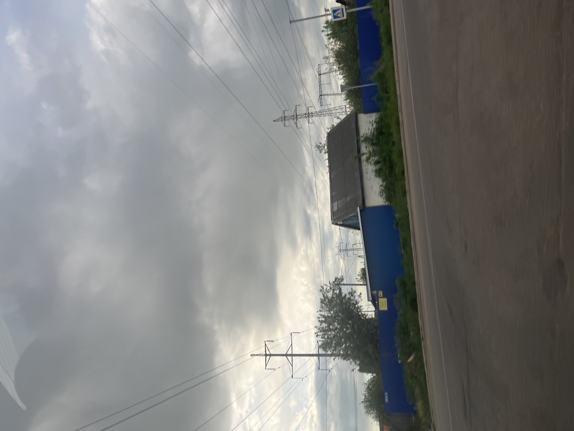 The image shows a street in Chaikovsky, Russia, beside a road with an asphalt surface. Power lines extend across the overcast sky. A blue fence parallels the road, interspersed with green vegetation. Buildings appear behind the fence, and road signs are visible.