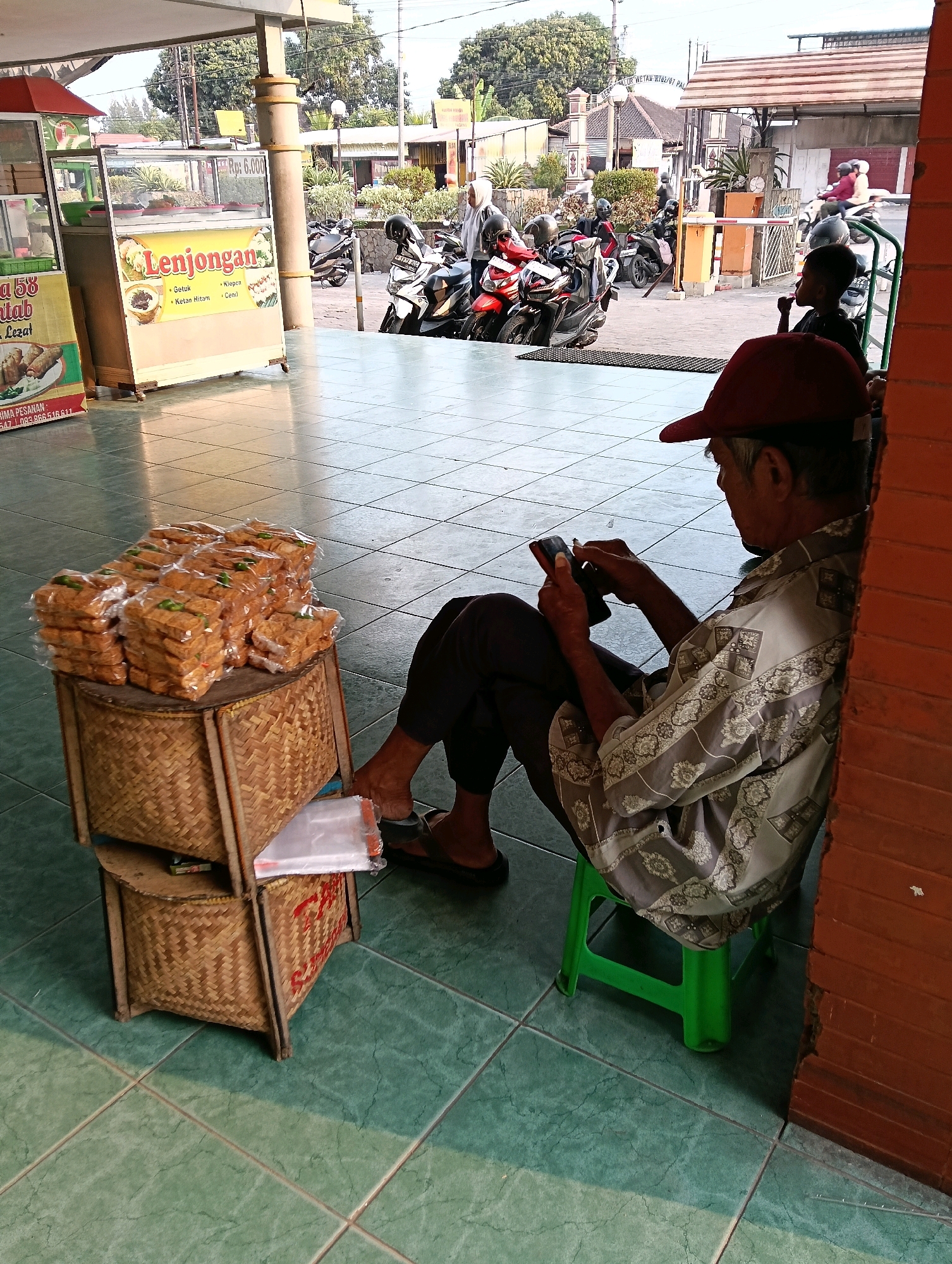 In Jaten, Indonesia, a street scene unfolds with a focus on a vendor and his merchandise. The foreground is dominated by a stack of neatly packaged, fried snacks, likely tempeh, arranged on a woven bamboo display. These are placed on a two-tiered woven bamboo stand, with a white plastic bag visible on the lower level. Beside the snacks, a man sits on a bright green plastic stool, leaning against a textured orange brick wall. He is wearing a red baseball cap and a patterned gray and white shirt, with dark pants and dark sandals. His attention is directed towards a black smartphone he holds in his hands. In the background, a food stall with a glass display case is visible, showcasing more food items and advertising "Lenjongan" with various prices and dishes. Motorbikes are parked in front of the stall, and several people, some wearing hijabs, are seen in the distance. The ground is tiled with a green checkered pattern, and the overall atmosphere suggests a busy marketplace or roadside eatery. The lighting indicates it is daytime, with sunlight casting shadows across the tiled floor.