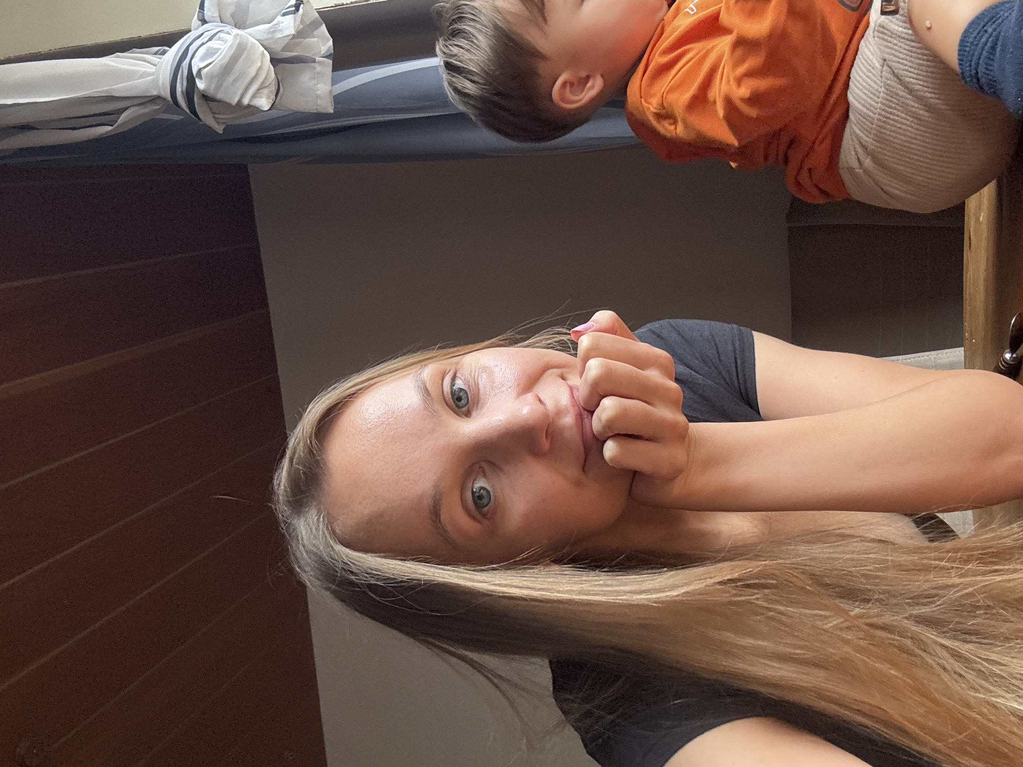 In a brightly lit room, a woman with long blonde hair rests her chin on her hand, her eyes looking towards the camera. To her right, a young child, facing away, is dressed in an orange shirt and light-colored pants. A curtain with a striped tieback hangs in the background, with the wooden paneled ceiling visible above. The scene appears to be indoors, likely a domestic setting within the city of Überherrn, Germany.