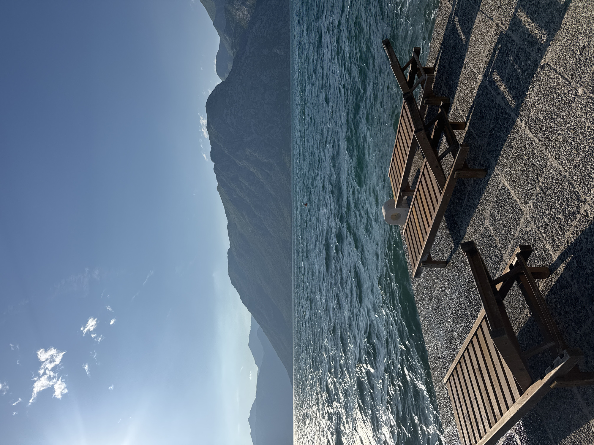 The image depicts a waterfront scene in Dobrota, Montenegro. Two wooden deck chairs sit on a paved walkway alongside the water. The water itself appears choppy, with sunlight reflecting on its surface. In the background, a mountain range is visible under a clear, blue sky with a few scattered clouds. The angle of the sun creates harsh shadows from the deck chairs on the pavement.