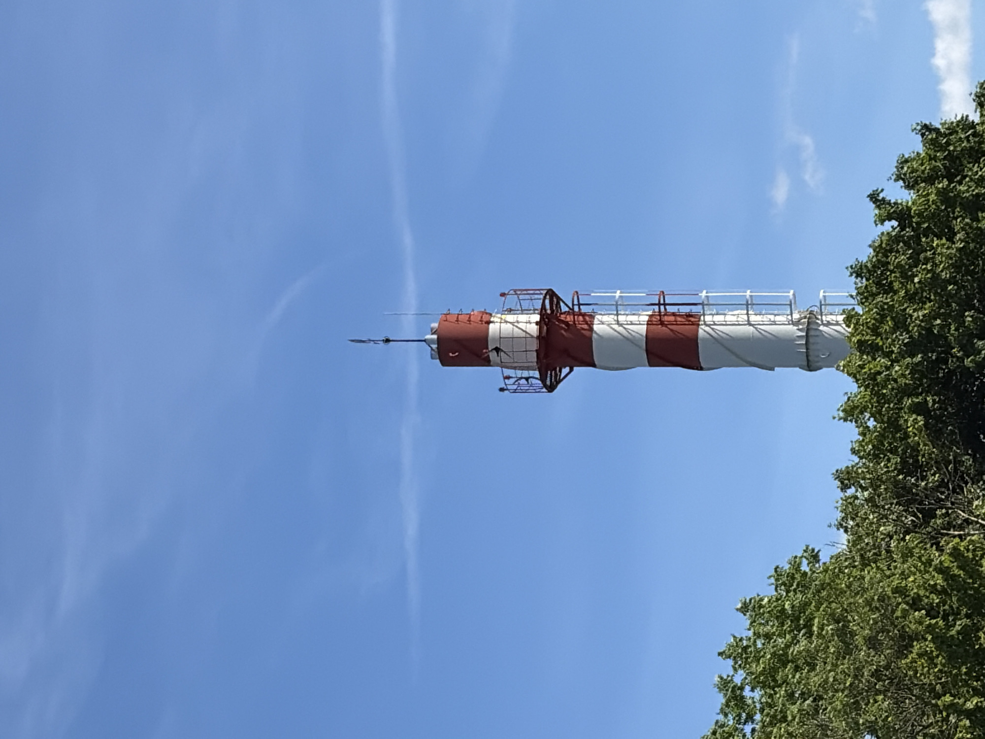 This is a low-angle outdoor shot of a tall, red and white striped structure against a bright blue sky. The structure appears to be a weather vane or a similar meteorological instrument, characterized by its cylindrical shape and a pointed spire at the top. It has a white railing around a section of it. To the right of the structure, the vibrant green foliage of a large tree is visible, its leaves forming a dense canopy. The sky is clear with only a few wispy white clouds, suggesting a sunny day. The orientation of the image is rotated, with the sky occupying the majority of the frame and the structure extending horizontally across the mid-section. There is no visible text in the image.