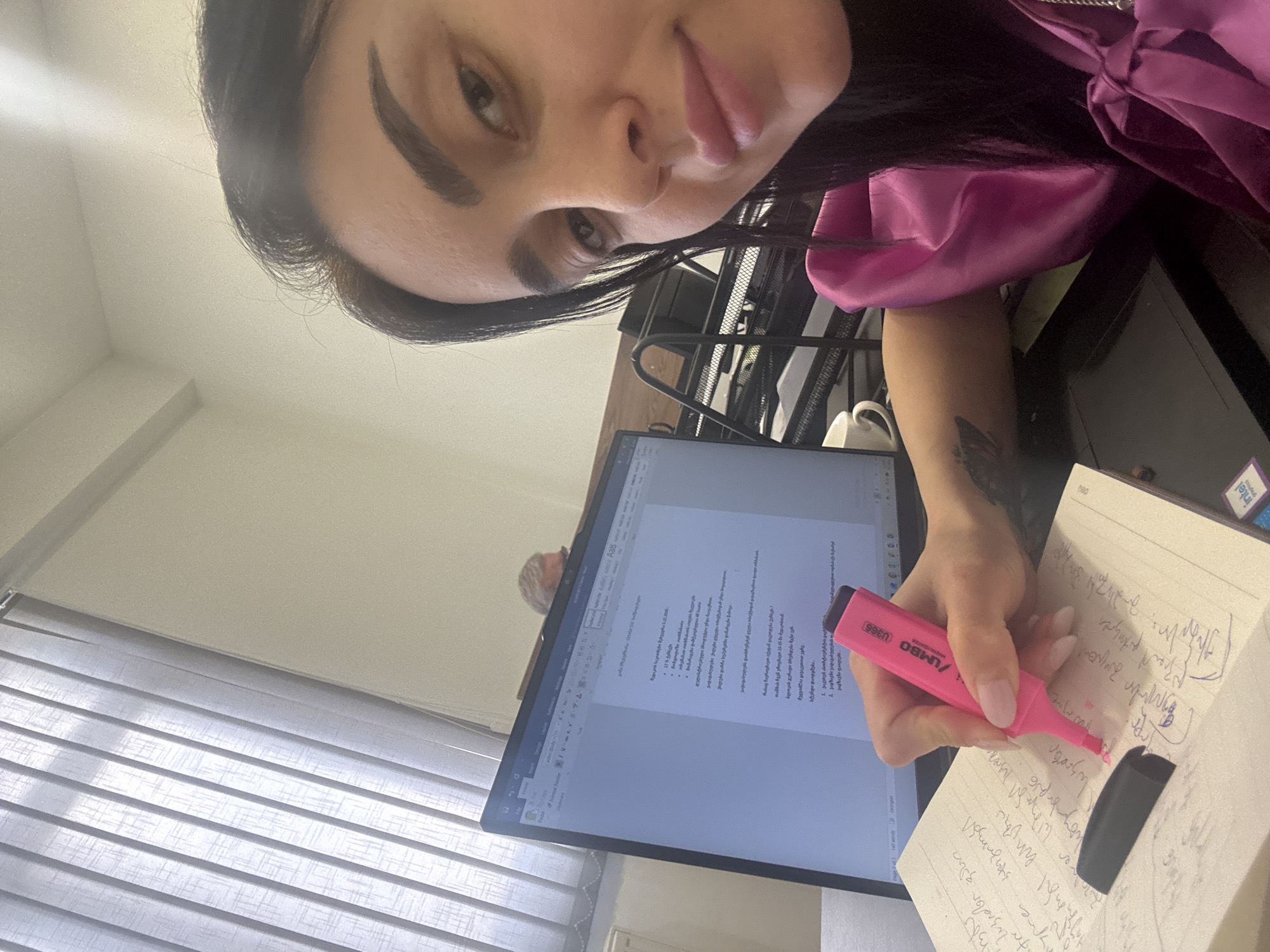 A woman is seated at a desk, working. She is highlighting notes in a notebook with a pink highlighter. A computer monitor displays a document with text written in Georgian and Persian. The desk is cluttered with various items, including a mug, an Intel sticker, and stationery. The setting appears to be an office or study space, possibly in Tbilisi, Georgia, given the presence of the Georgian language in the document on the screen. The woman is wearing a bright pink top. The window is covered by blinds. The woman has dark hair and dark eyebrows.